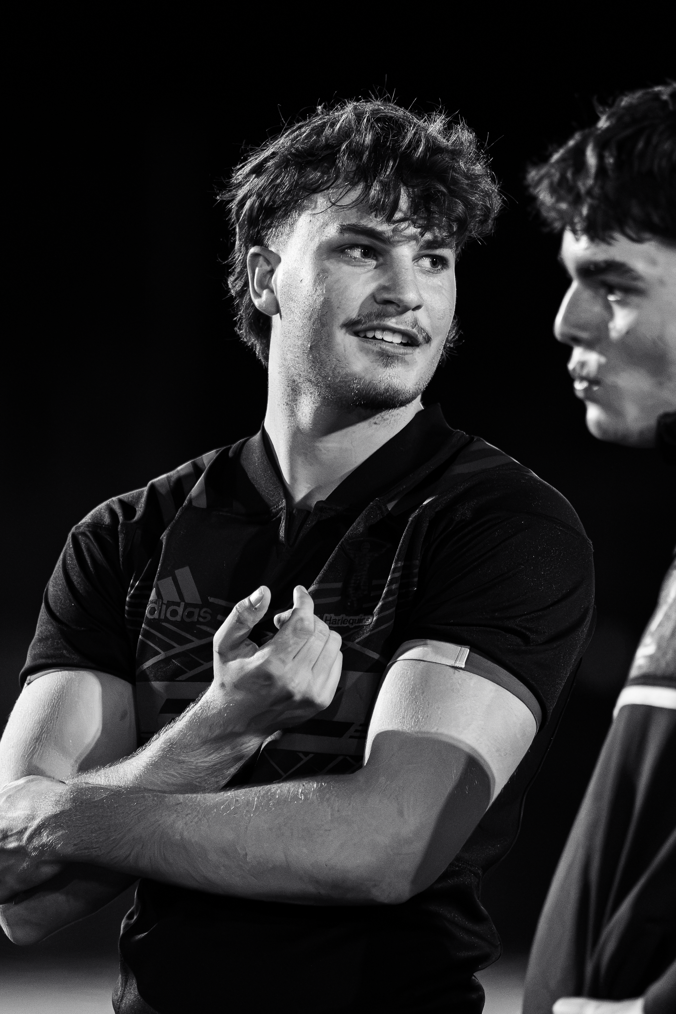 Two young men, one with curly hair, are engaging in a conversation, possibly during a sports event. The man on the left is smiling and resting his arm on a surface, while the other is looking at him. They are wearing athletic clothing.