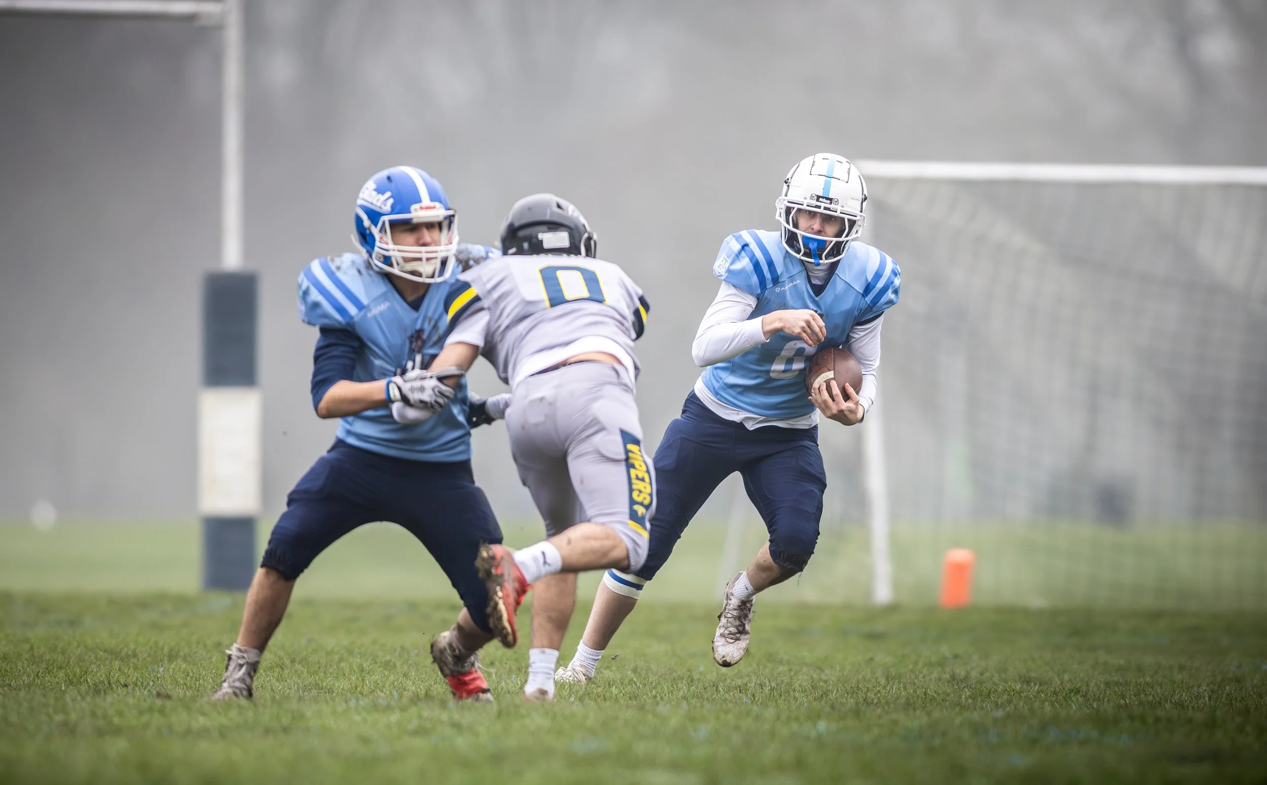 A youth football game with players in blue and gray uniforms on a grassy field, with one player holding the football and running, while others attempt to block or tackle.