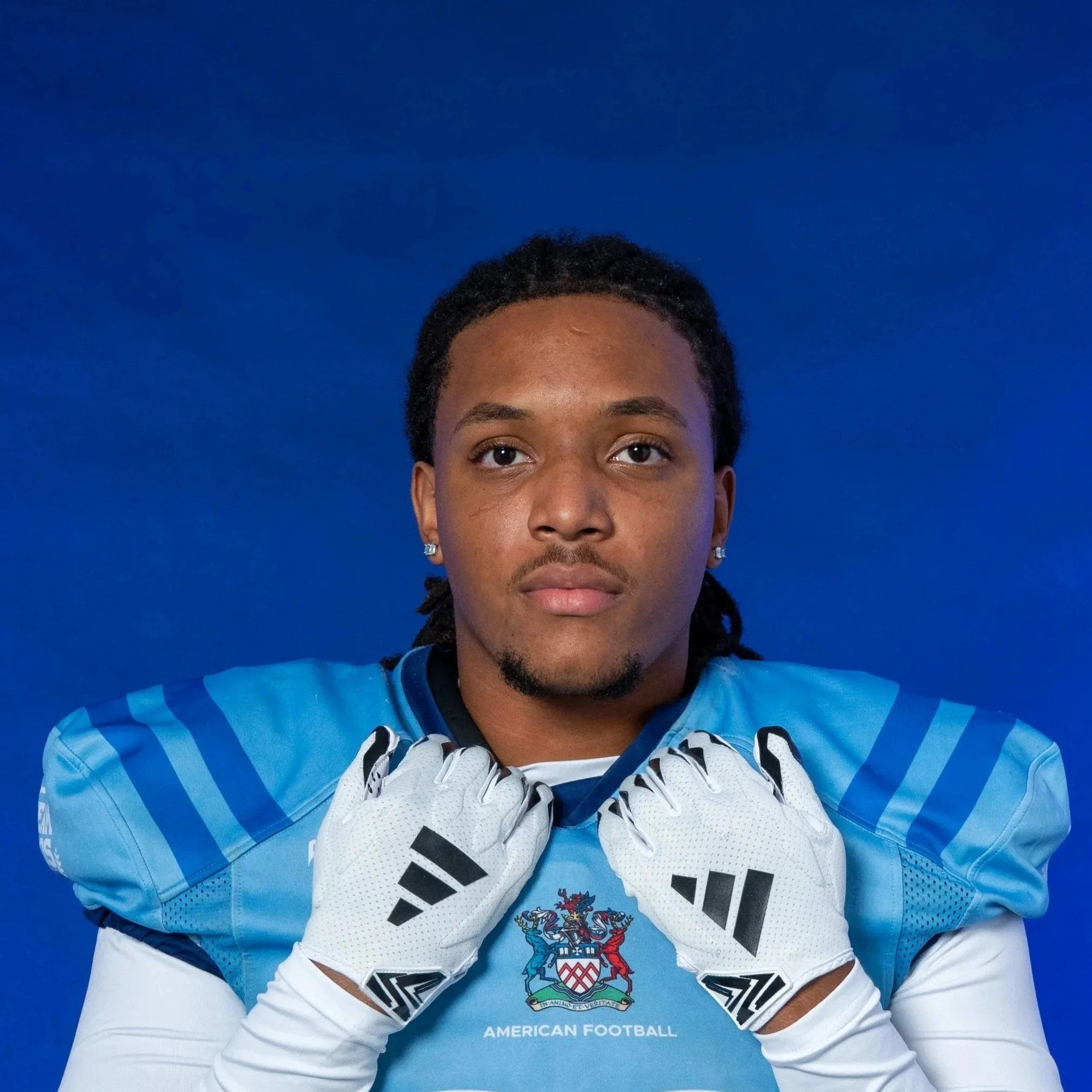A football player wearing a blue helmet with white stripes, red gloves, and a blue jersey with shoulder padding. The player is holding the chin strap of his helmet with both hands and looking directly at the camera. The background is dark.