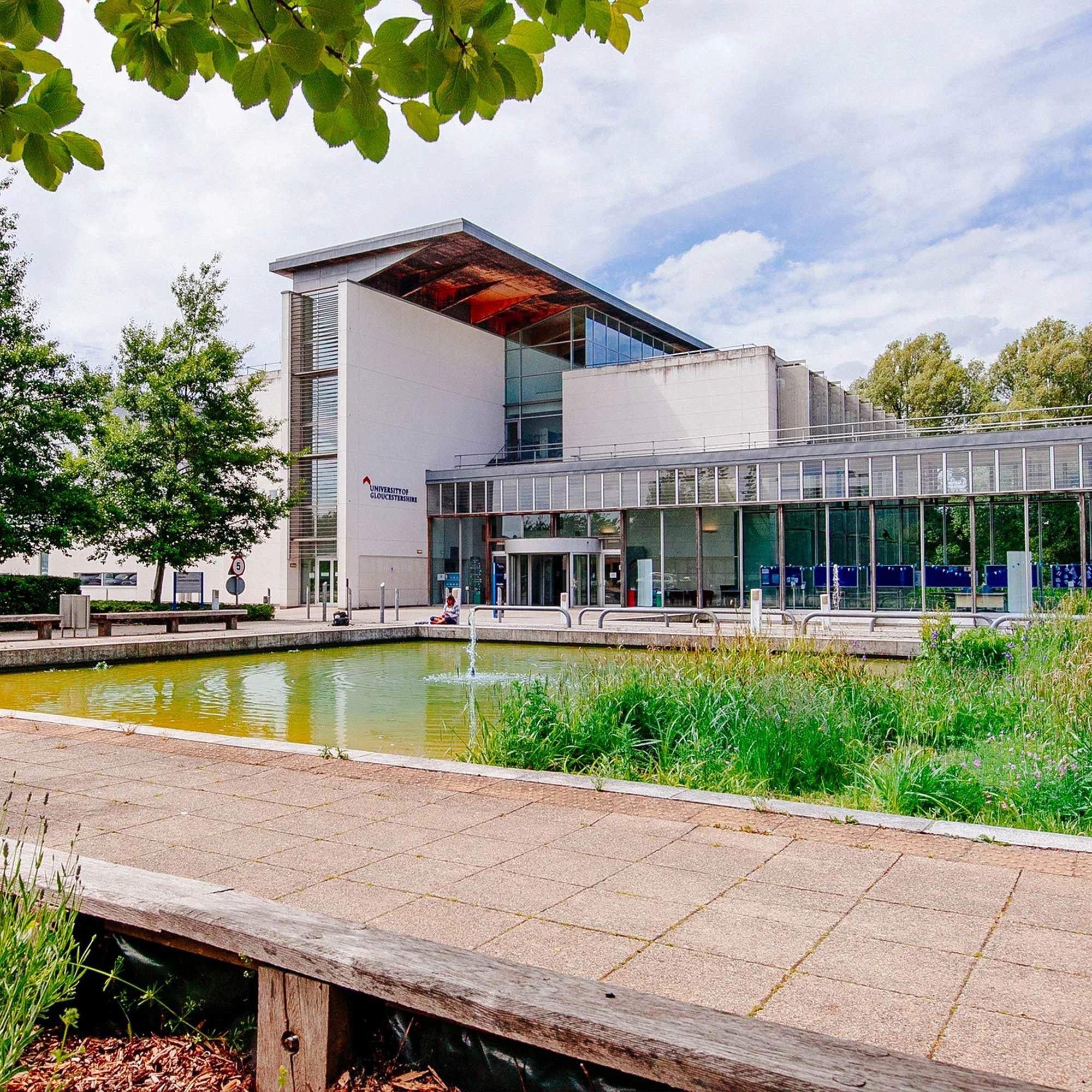 Modern university building with glass and white exterior, a water pond in the foreground, surrounded by greenery and trees, under a partly cloudy sky.