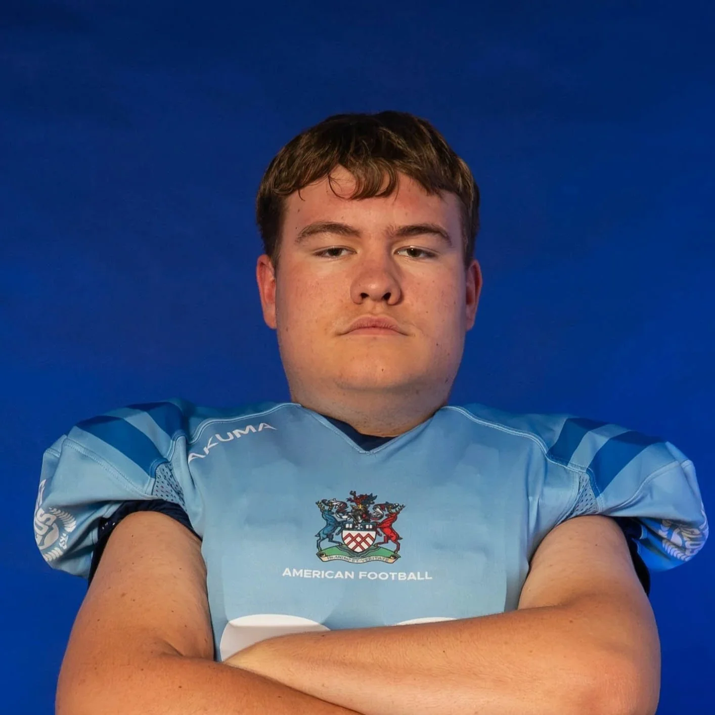 A football player wearing a blue helmet and jersey, holding a football, standing in front of a blue background with the GLOWS logo.