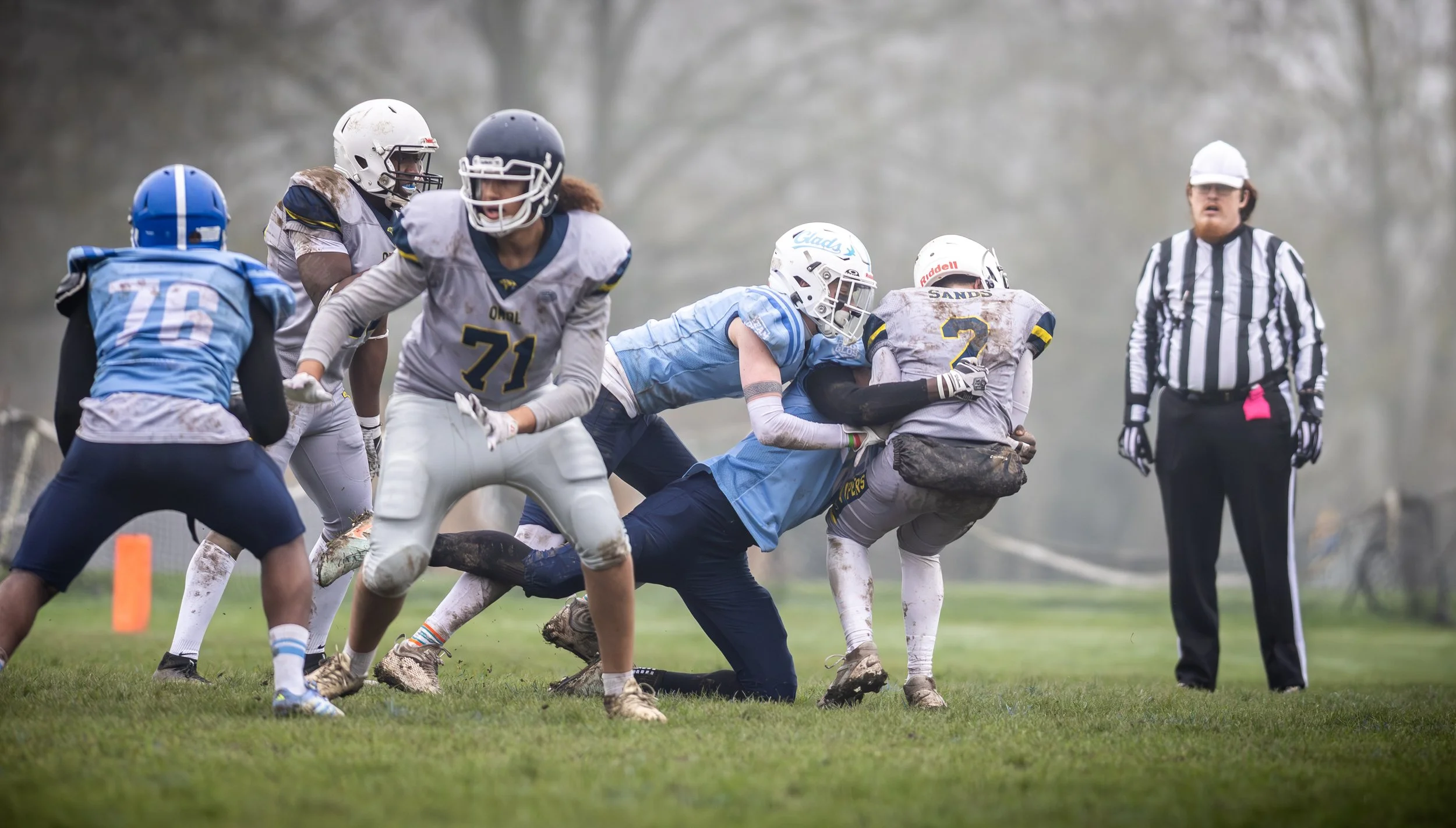 A group of youth football players on a muddy field, with one player tackling another, and a referee observing in the background.