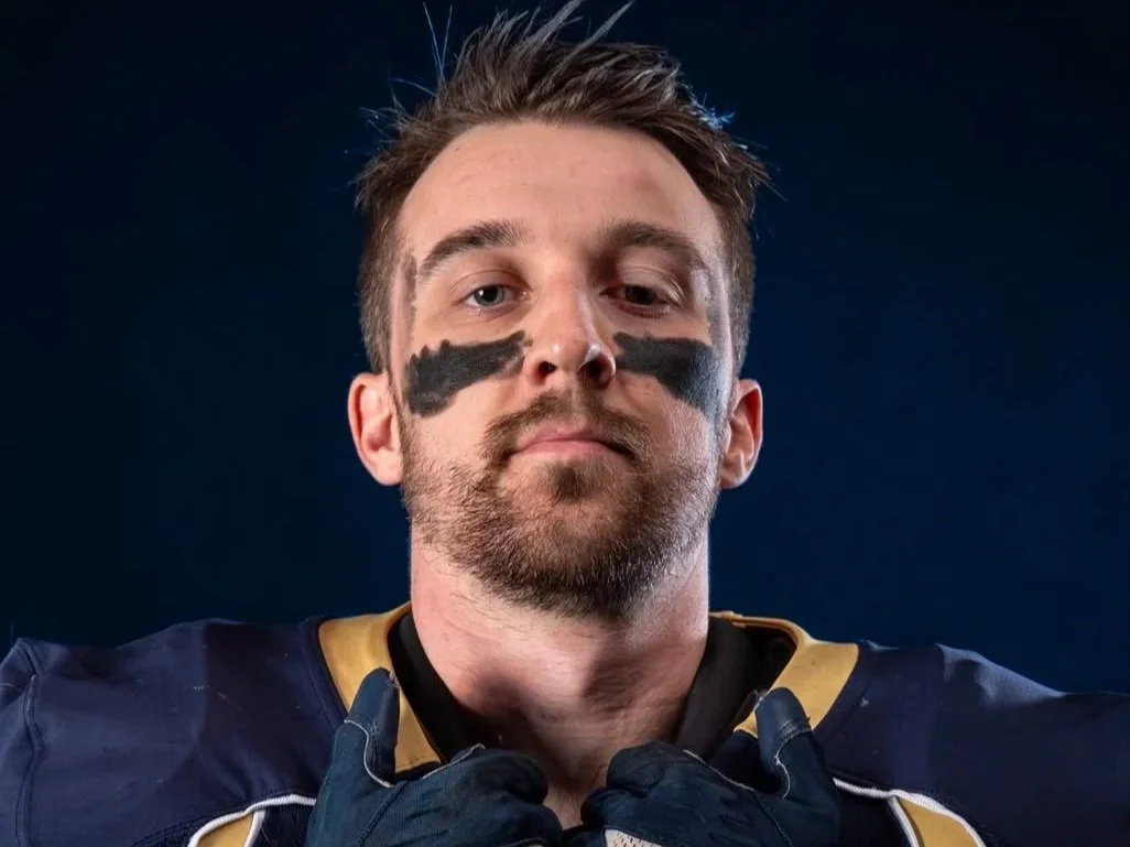 A football player with face paint, wearing a dark uniform with gold accents, holding his collar.