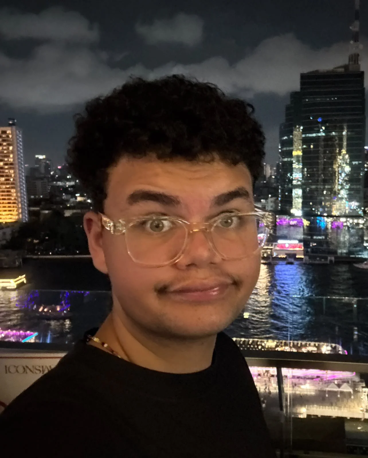 A young man with glasses and curly hair taking a selfie at night with a city skyline and water in the background.