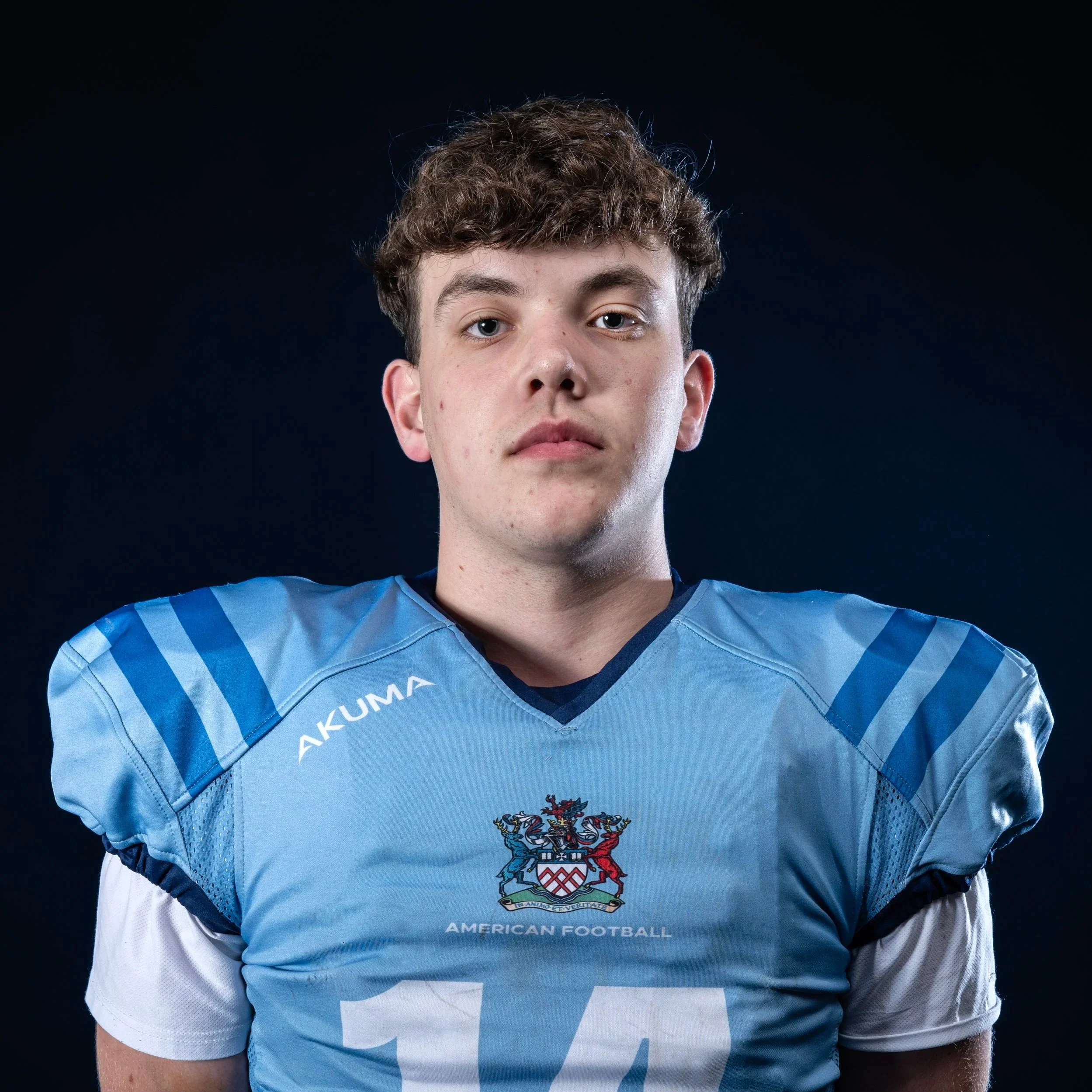 Close-up of a young male athlete wearing a blue sports jersey with crest emblem, short brown hair, and a neutral expression, against a dark background.