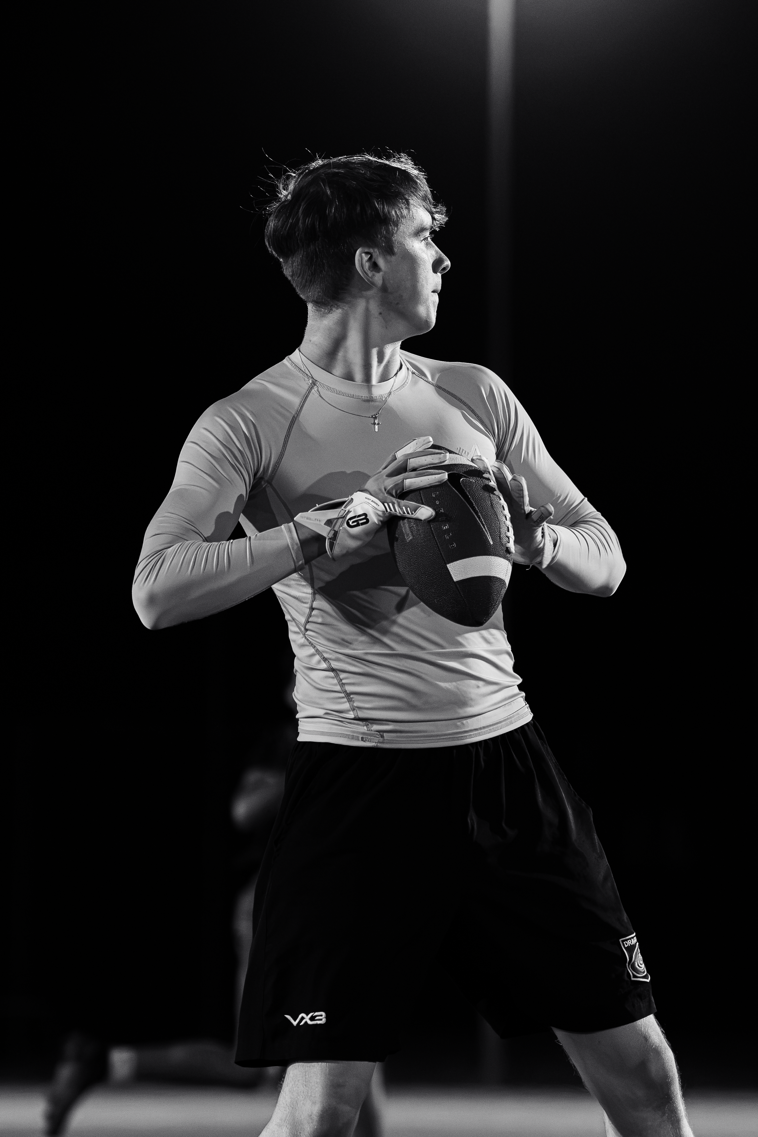 A young male athlete with short hair, wearing sports gloves, a T-shirt, and shorts holding a football in a ready stance during a game or practice.