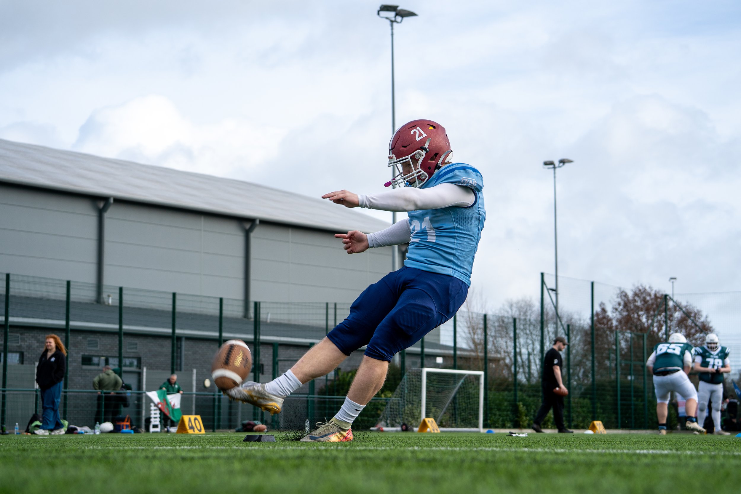 A football player in a blue uniform and red helmet is kicking a football during practice on an outdoor field, with other players and coaches in the background.