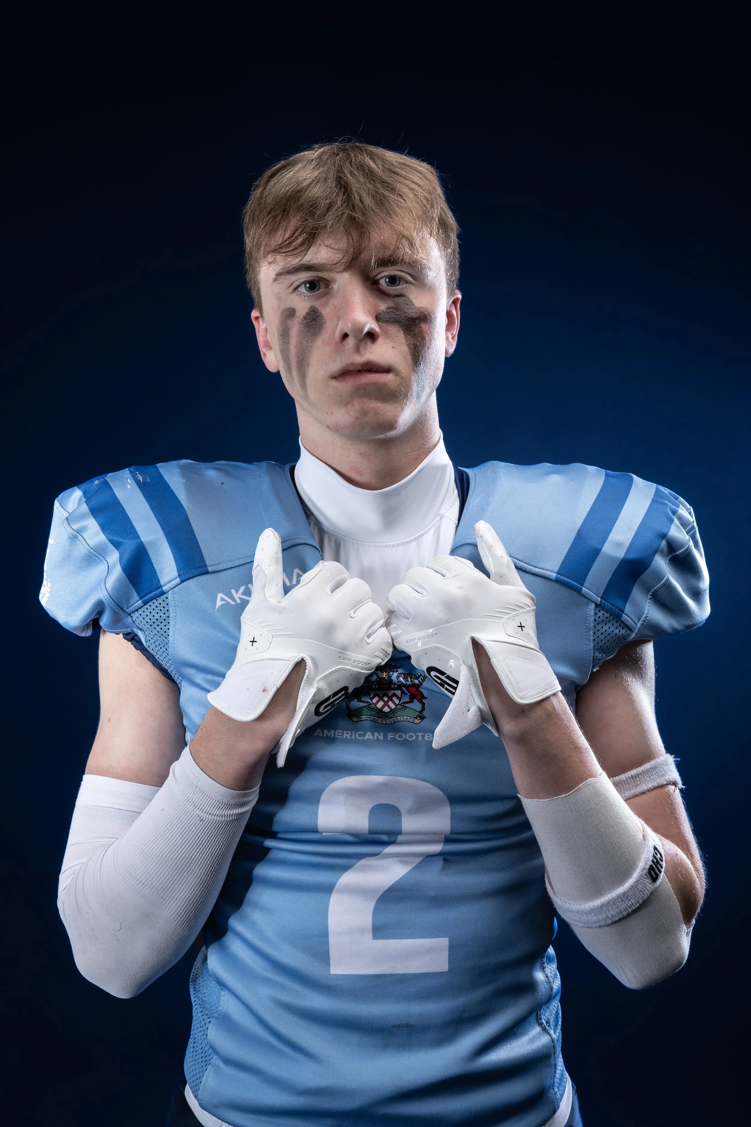 A young male football player with short reddish-brown hair wearing a light blue football jersey with the number 2, white gloves, and white arm sleeves, standing against a dark background, looking directly at the camera.