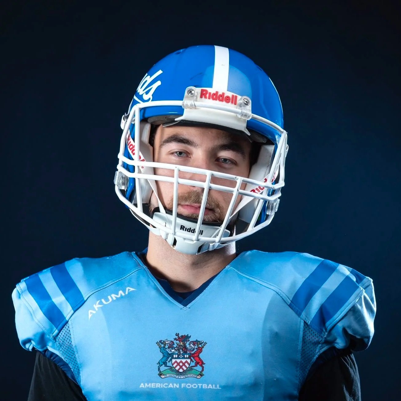 Portrait of a male football player wearing a blue helmet and jersey with an American football team crest, against a dark background.