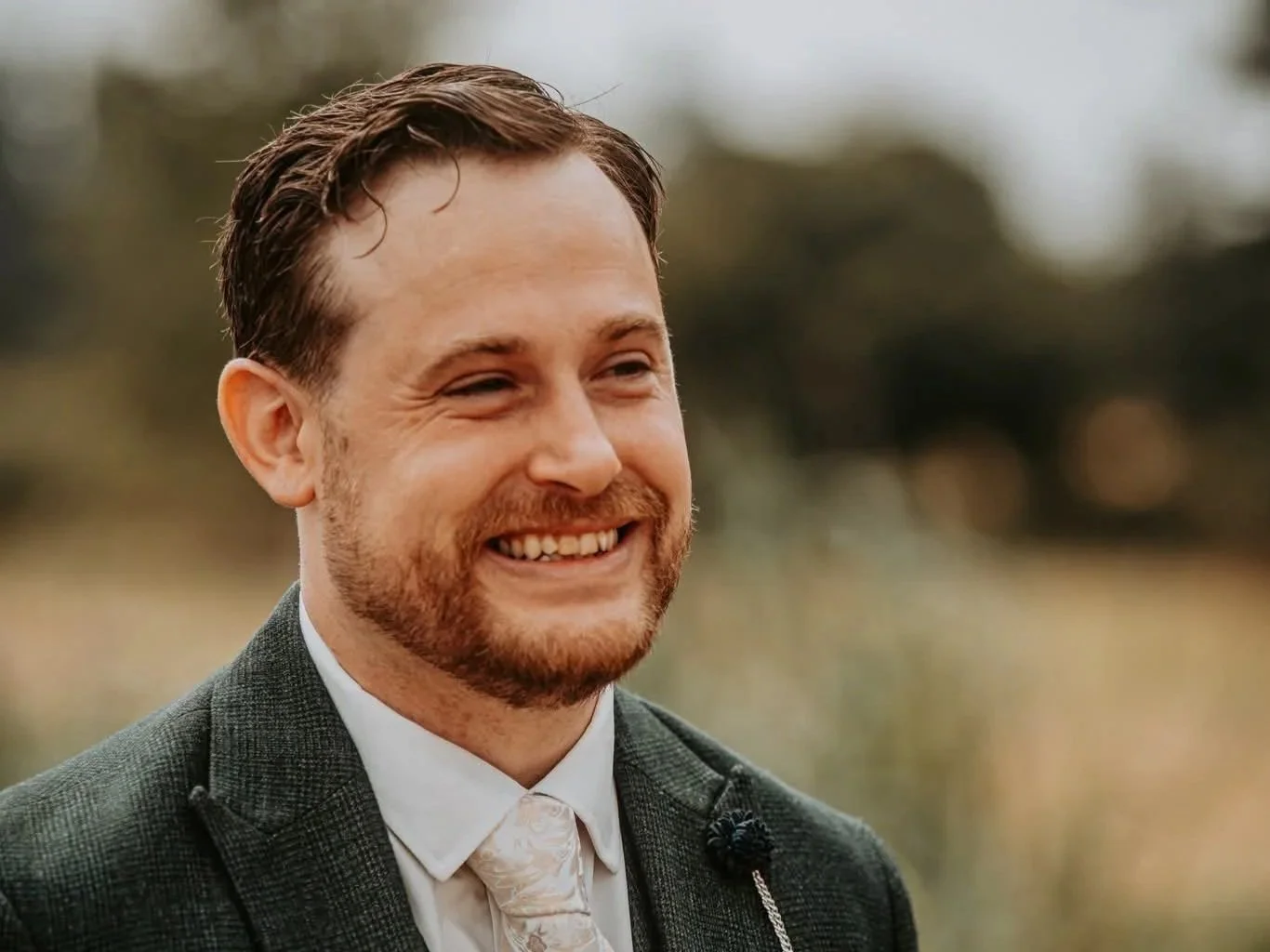 A smiling man with brown hair and beard, wearing a dark suit and white shirt, standing outdoors with a blurred background.