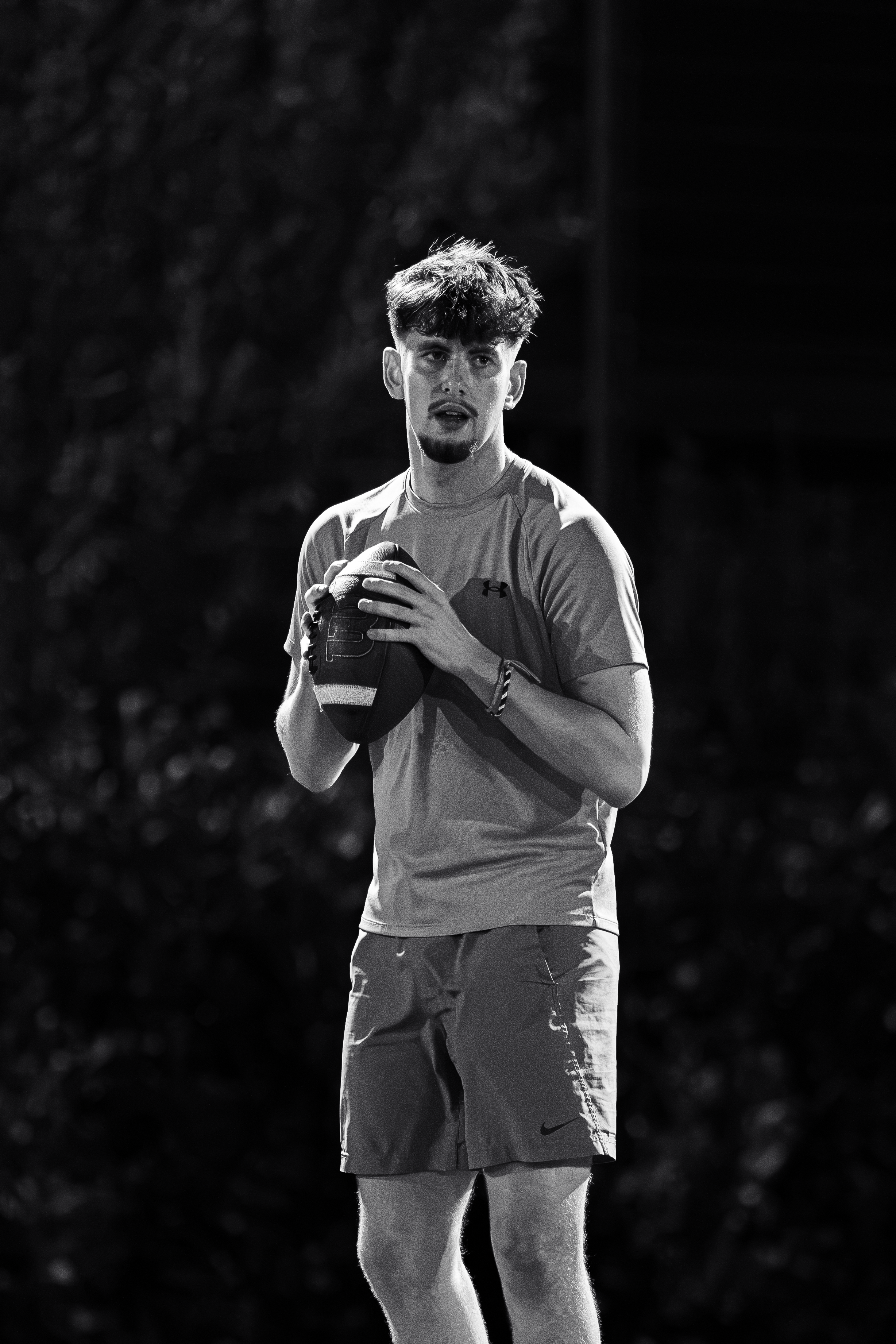A young man with dark, curly hair playing American football outdoors at night, wearing athletic clothing and holding a football.