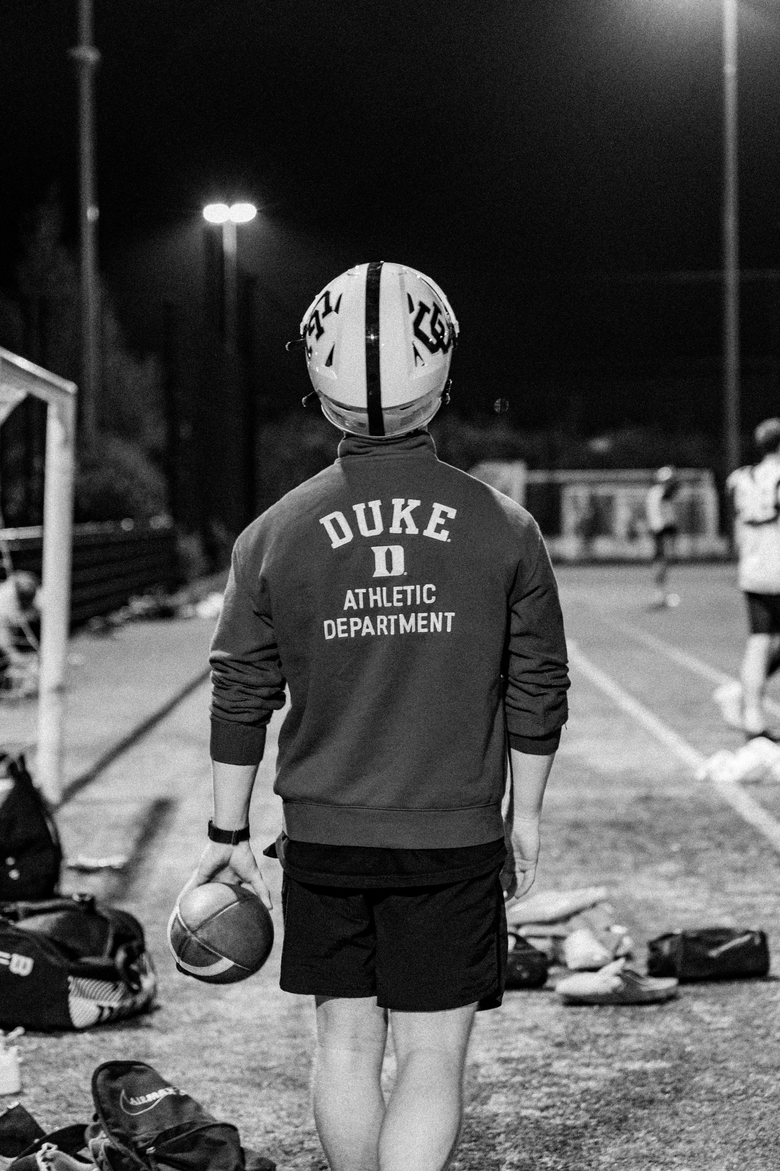 A person wearing a helmet and sweatshirt with 'Duke Athletic Department' on back, standing on a sports field at night holding a football, with other athletes and sports equipment in the background.