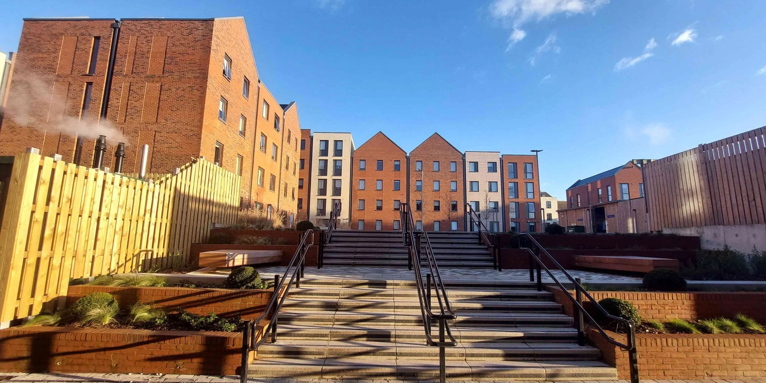 Urban apartment complex with stairs, brick and modern buildings, sunny sky, landscaped garden with plants, wooden fencing, and benches.