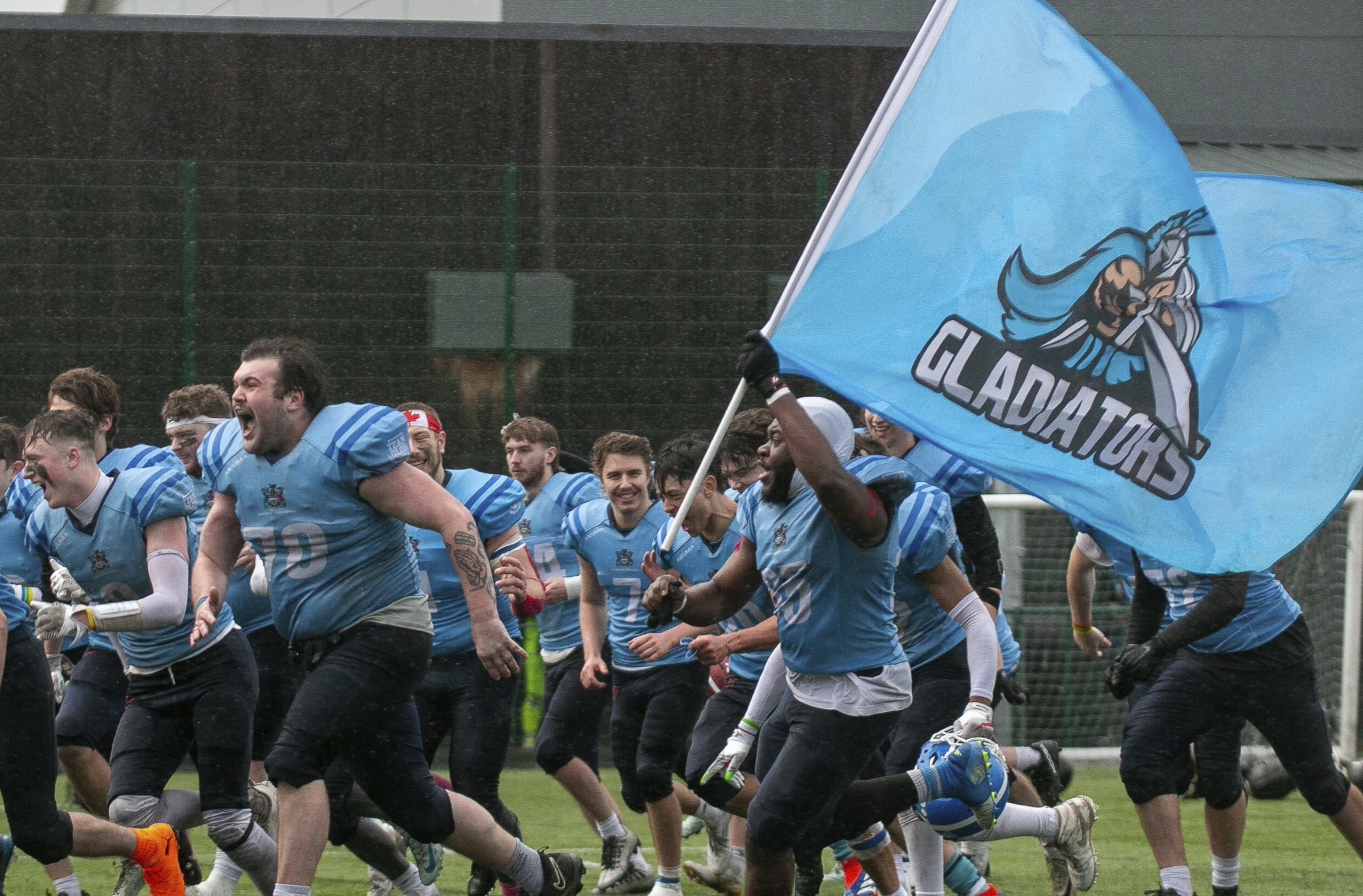 A group of American football players wearing blue jerseys are running on a field during a game, with one player holding a flag with the team logo.