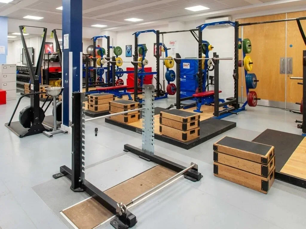 Empty gym with weightlifting equipment, including squat racks, barbells, weight plates, plyometric boxes, and a rowing machine.
