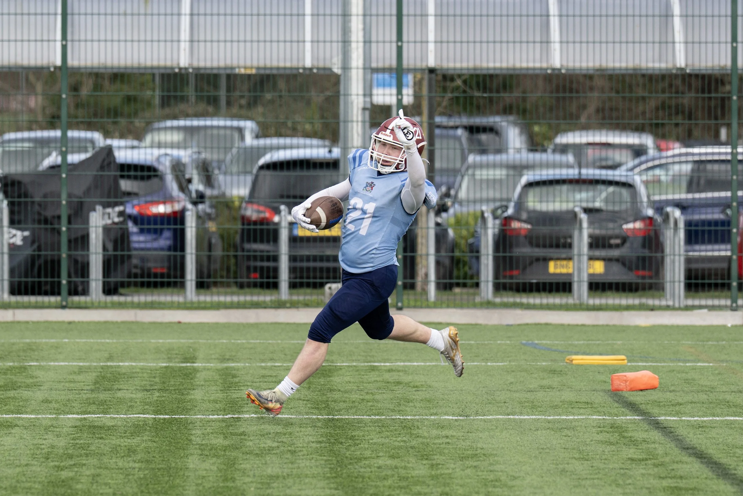 A young male football player wearing a blue jersey with number 21, a red helmet, white gloves, and running cleats is running on a sports field holding a football, with parking lot and cars visible behind a green fence.