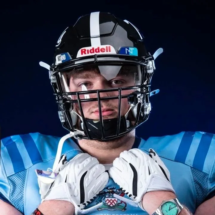 A football player wearing a black helmet with a faceguard and a blue jersey, standing against a dark background.