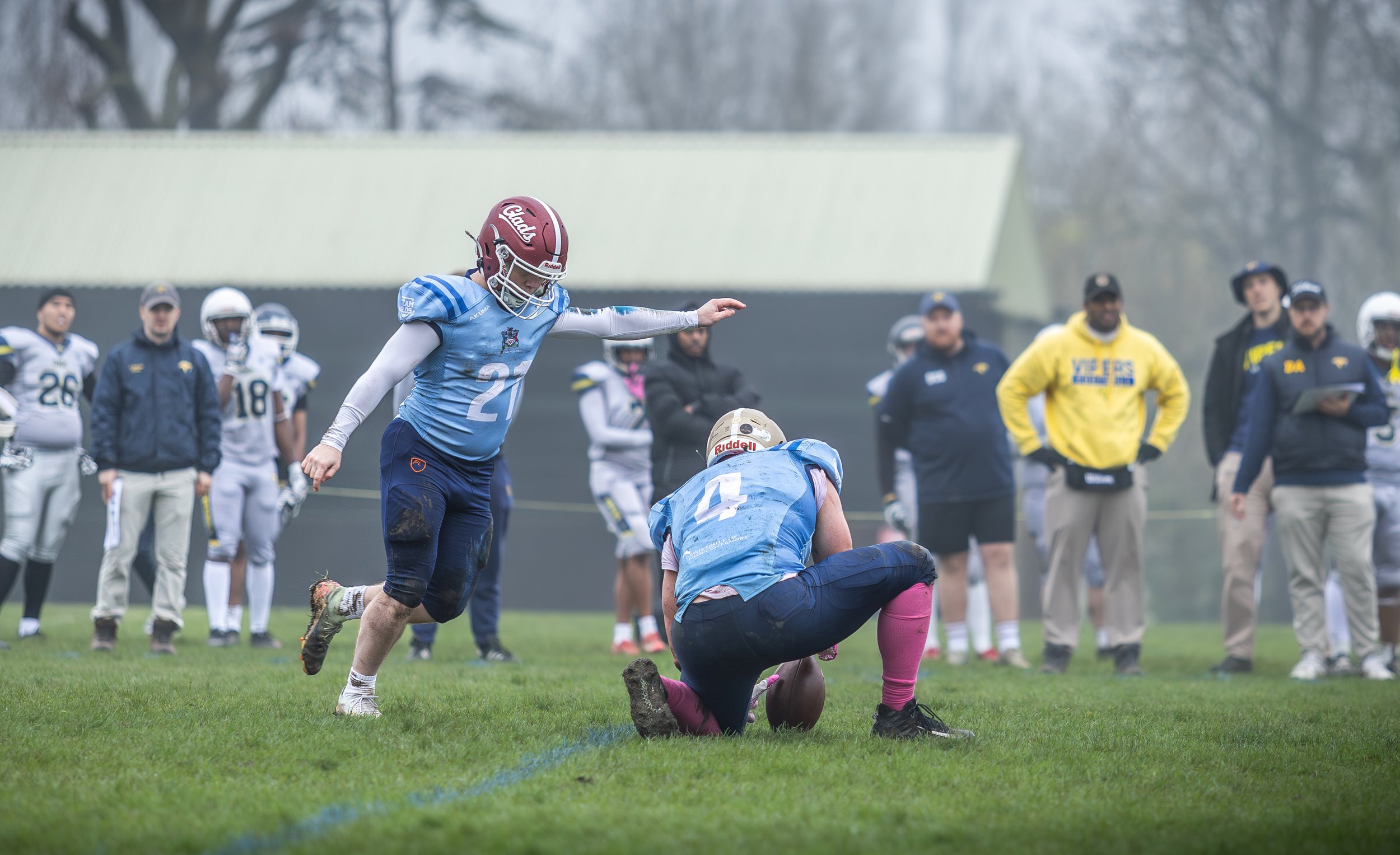 A football player in a blue jersey with the number 21 is tossing a football to a teammate on a grassy field. Several other players and spectators stand in the background, some wearing jackets and caps, on a foggy day.
