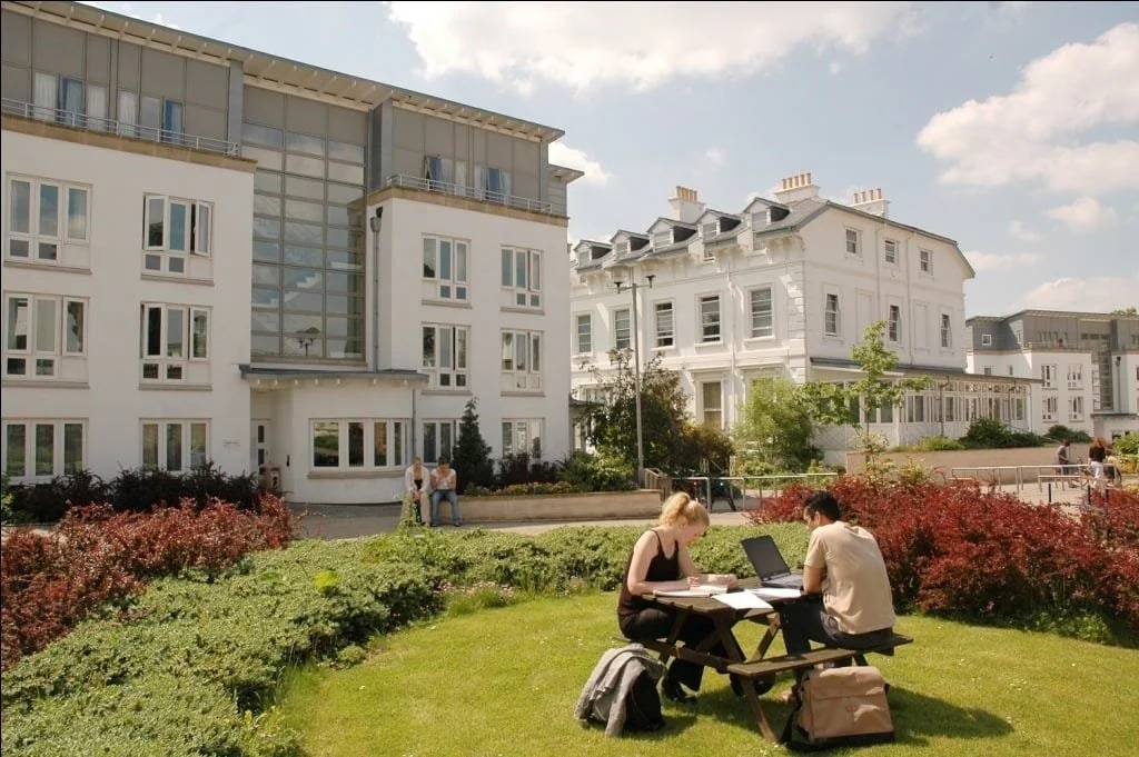 Two people working or studying at a picnic table outdoors on a grassy area, with modern white apartment buildings and a landscaped garden in the background.