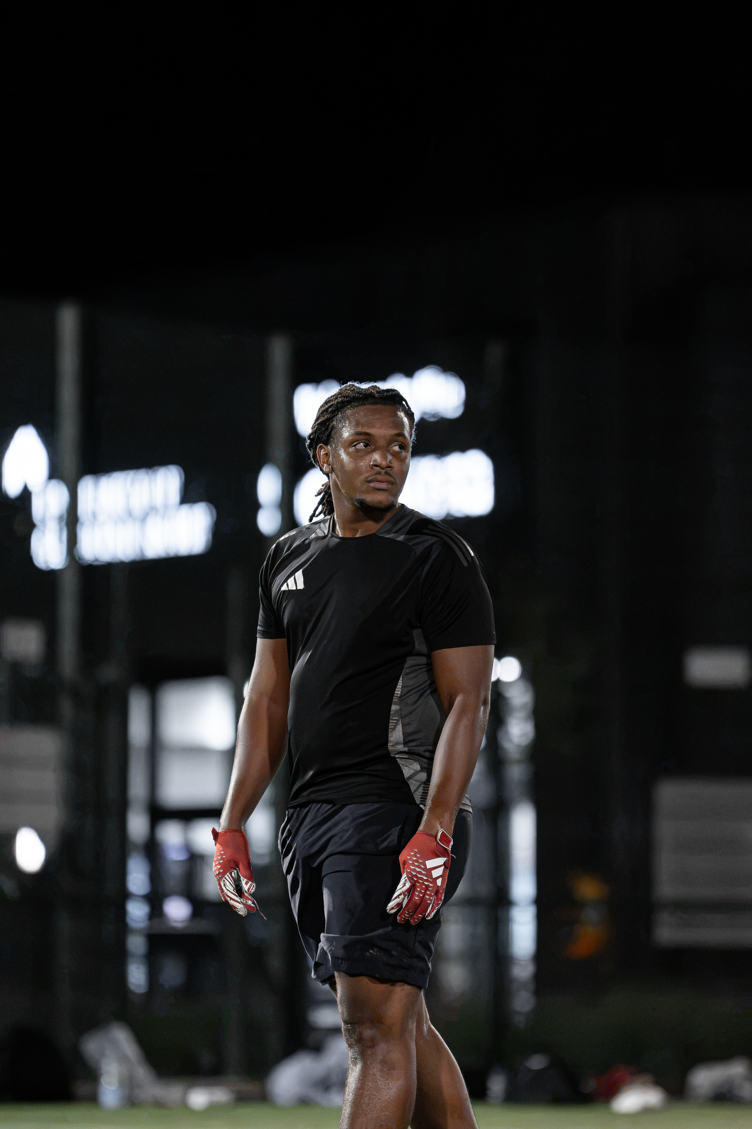A male athlete with dreadlocks on an indoor sports field, wearing a black athletic shirt, shorts, and red gloves, standing with a serious expression, with blurred indoor sports equipment in the background.