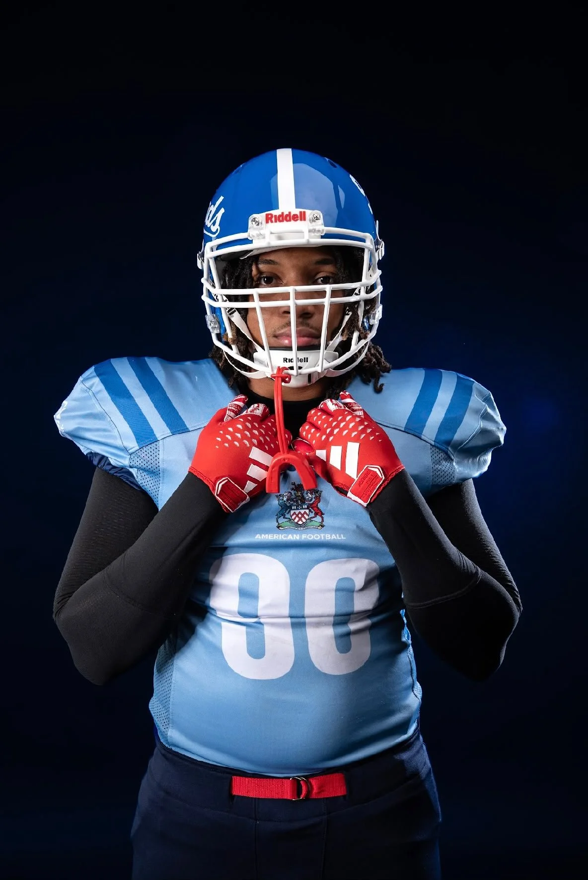Indigenous American football player wearing a blue jersey and helmet, holding the chin strap, posing against a black background.