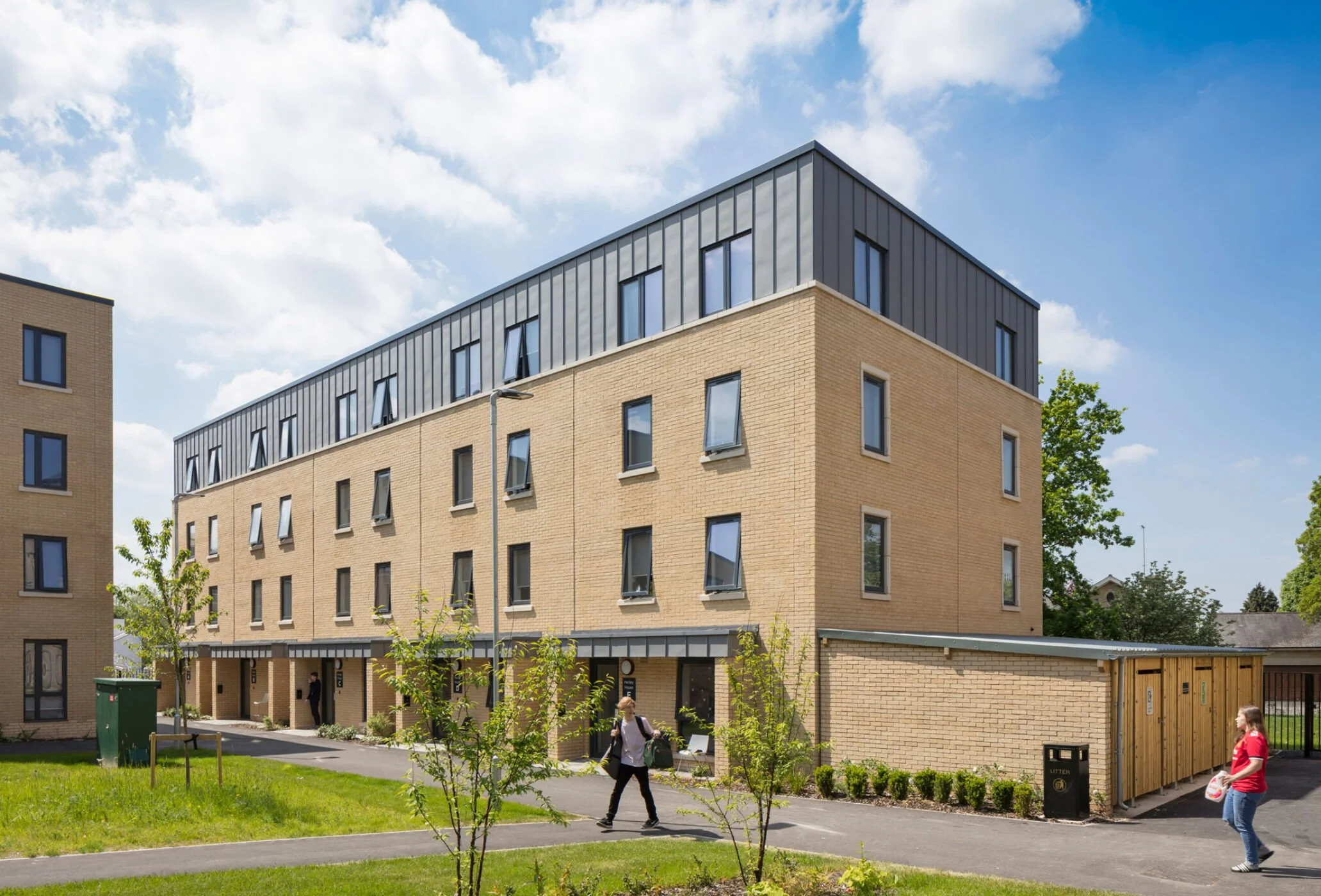 Modern brick apartment building with several windows and a black metal top floor, surrounded by trees and grass, with people walking outside on a sunny day.