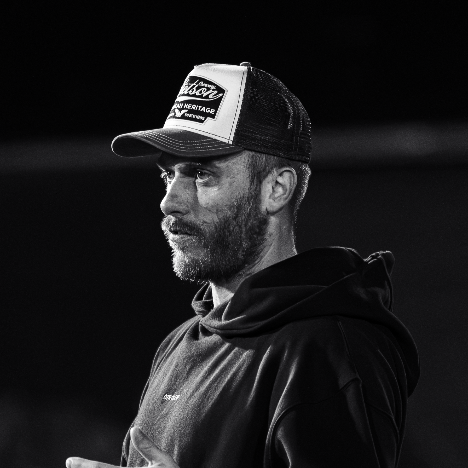 A black and white photograph of a man with a beard wearing a trucker hat and a hoodie, looking to the left with a contemplative expression.