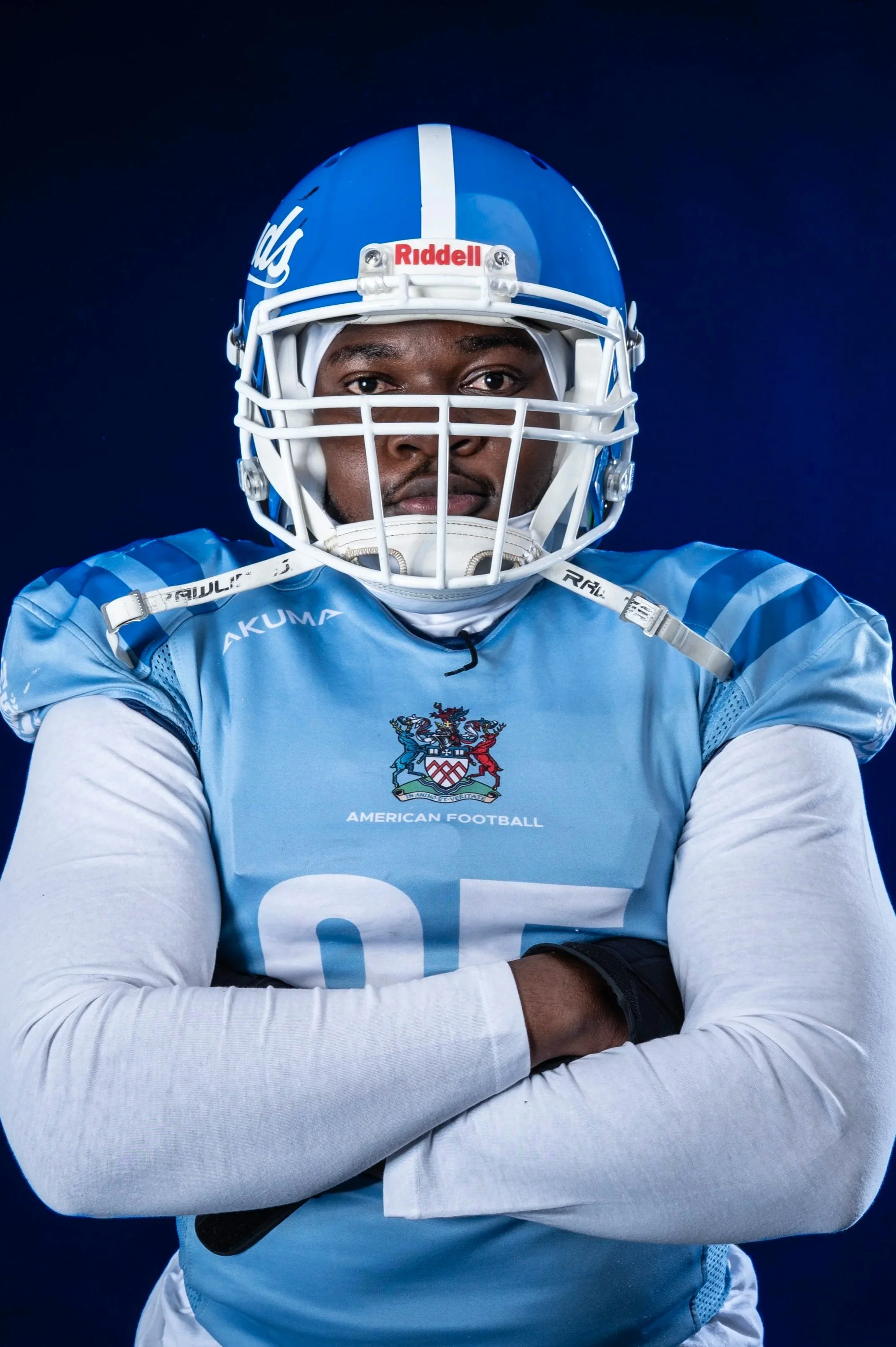 Portrait of a football player wearing a blue helmet and uniform with arms crossed, against a dark background.