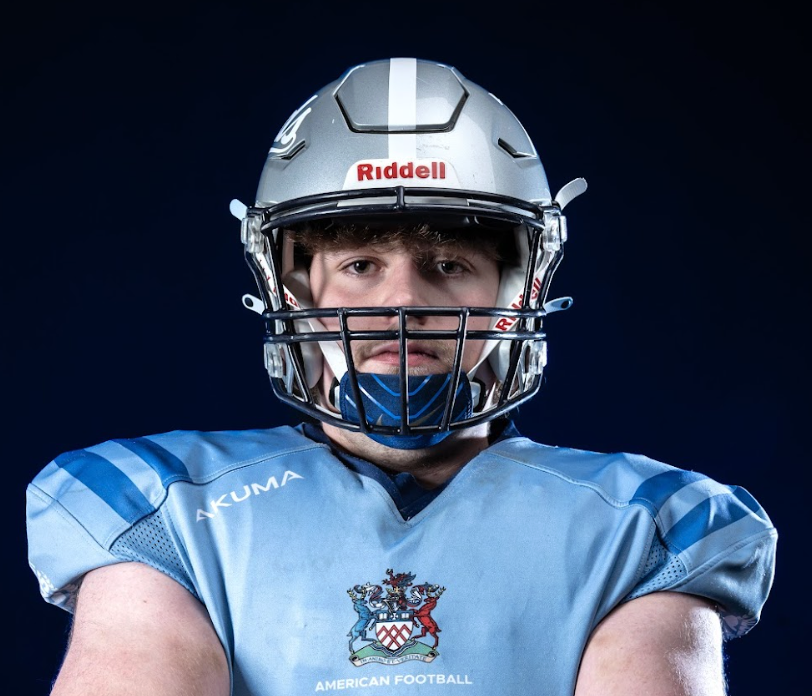 Portrait of a young male football player in a blue uniform and gray helmet with a faceguard, against a dark background.