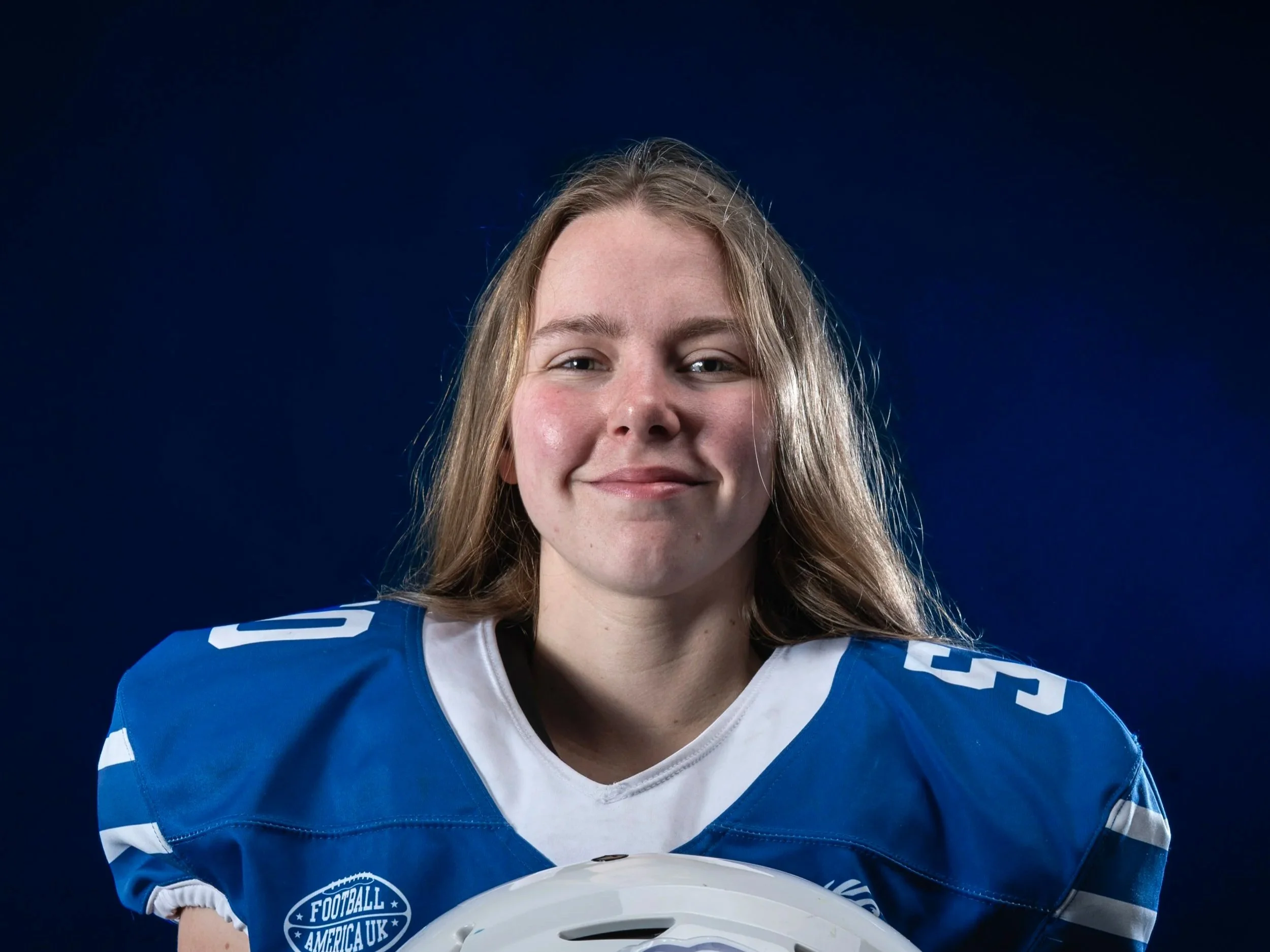 A young woman with light brown hair smiling wearing a blue and white sports jersey, holding a white helmet, against a dark background.