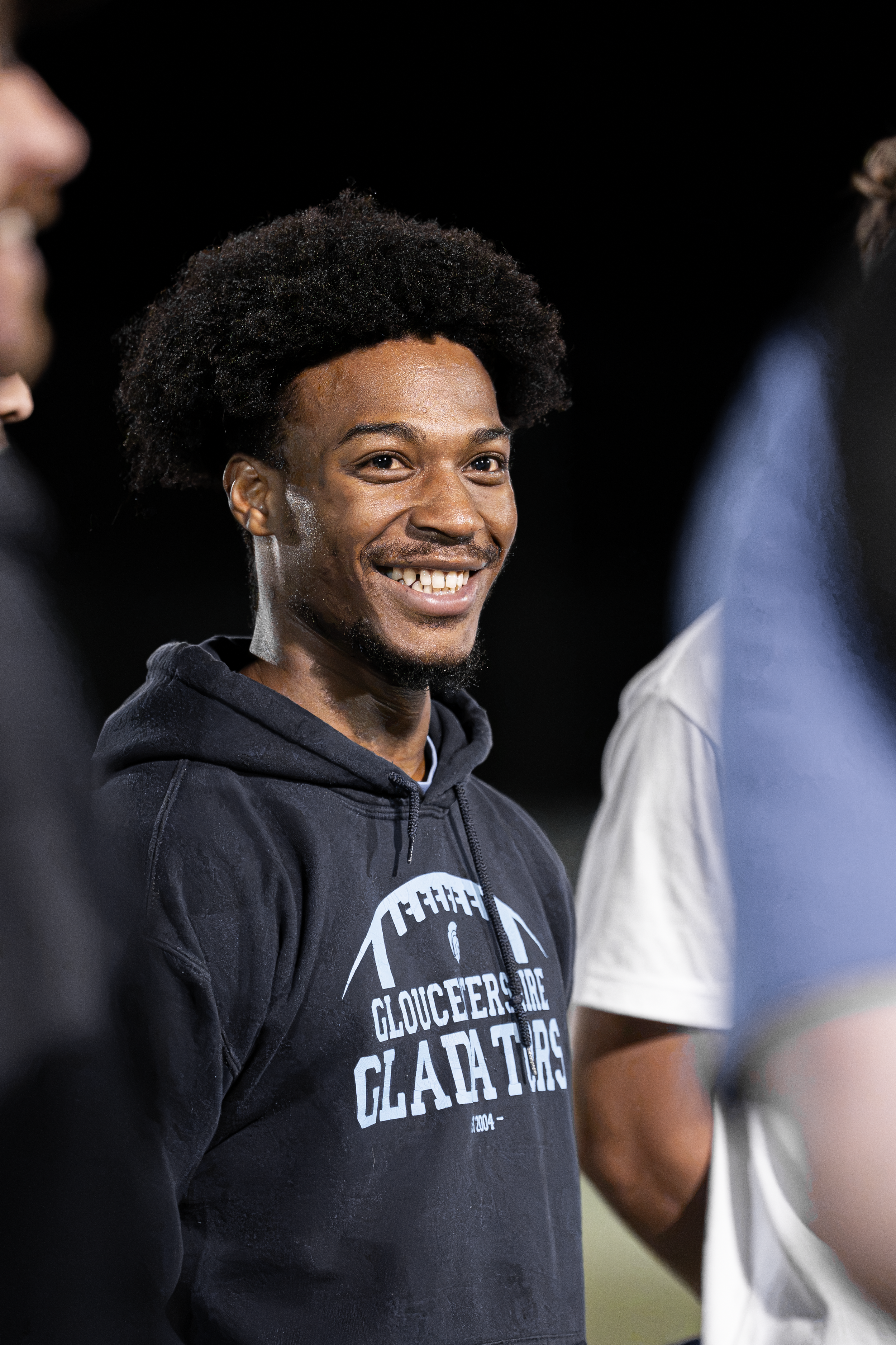 Smiling man with curly hair wearing a dark navy hoodie that says 'Gloucestershire Gladiators' standing between two people, partly visible, against a black background.