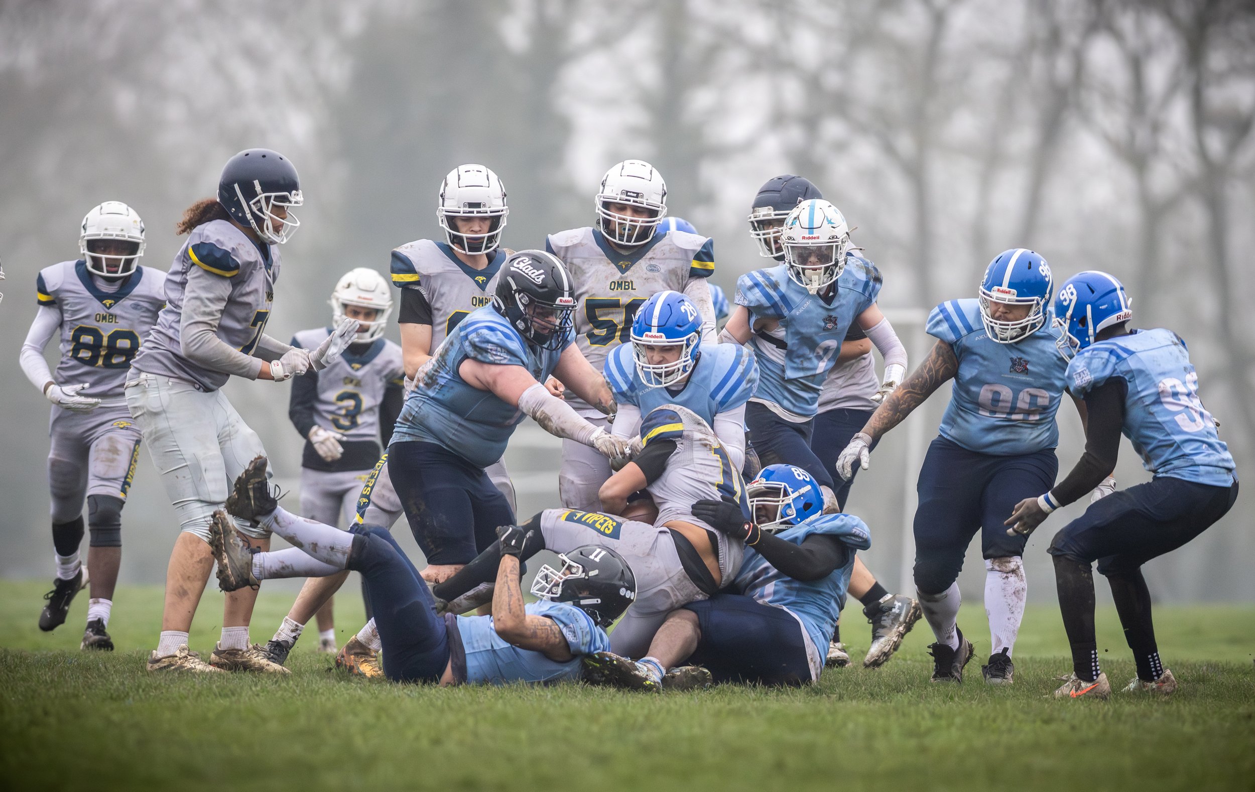 An American football game where players are engaged in a tackle on the field. Some players are wearing white and gray uniforms, while others are dressed in blue uniforms. Players are in various positions, with some standing and others on the ground, involved in the play, and the background shows blurred trees on a rainy day.