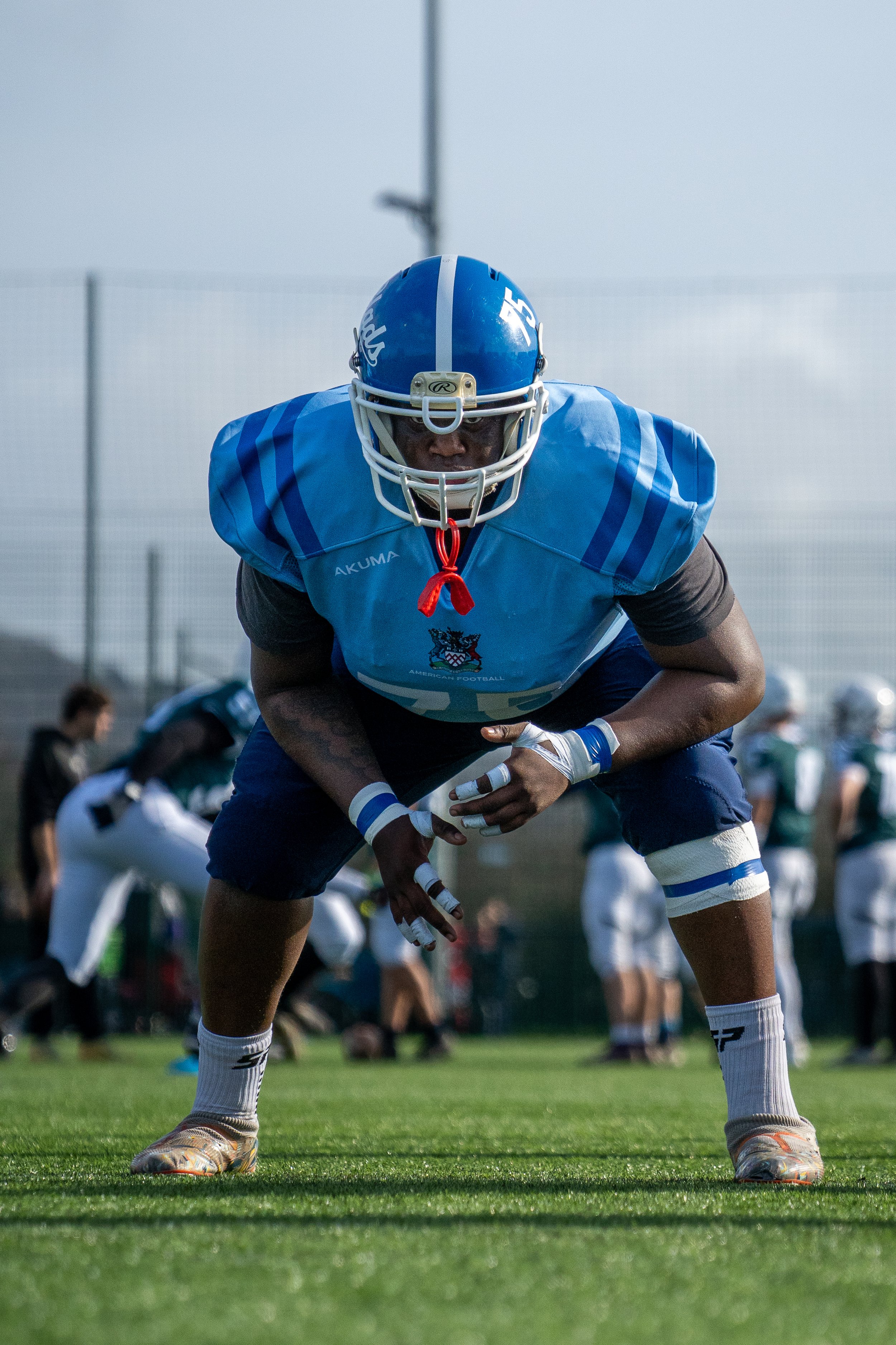 American football player in blue uniform crouching on field during practice or game, wearing helmet, gloves, and cleats, with other players blurred in background.