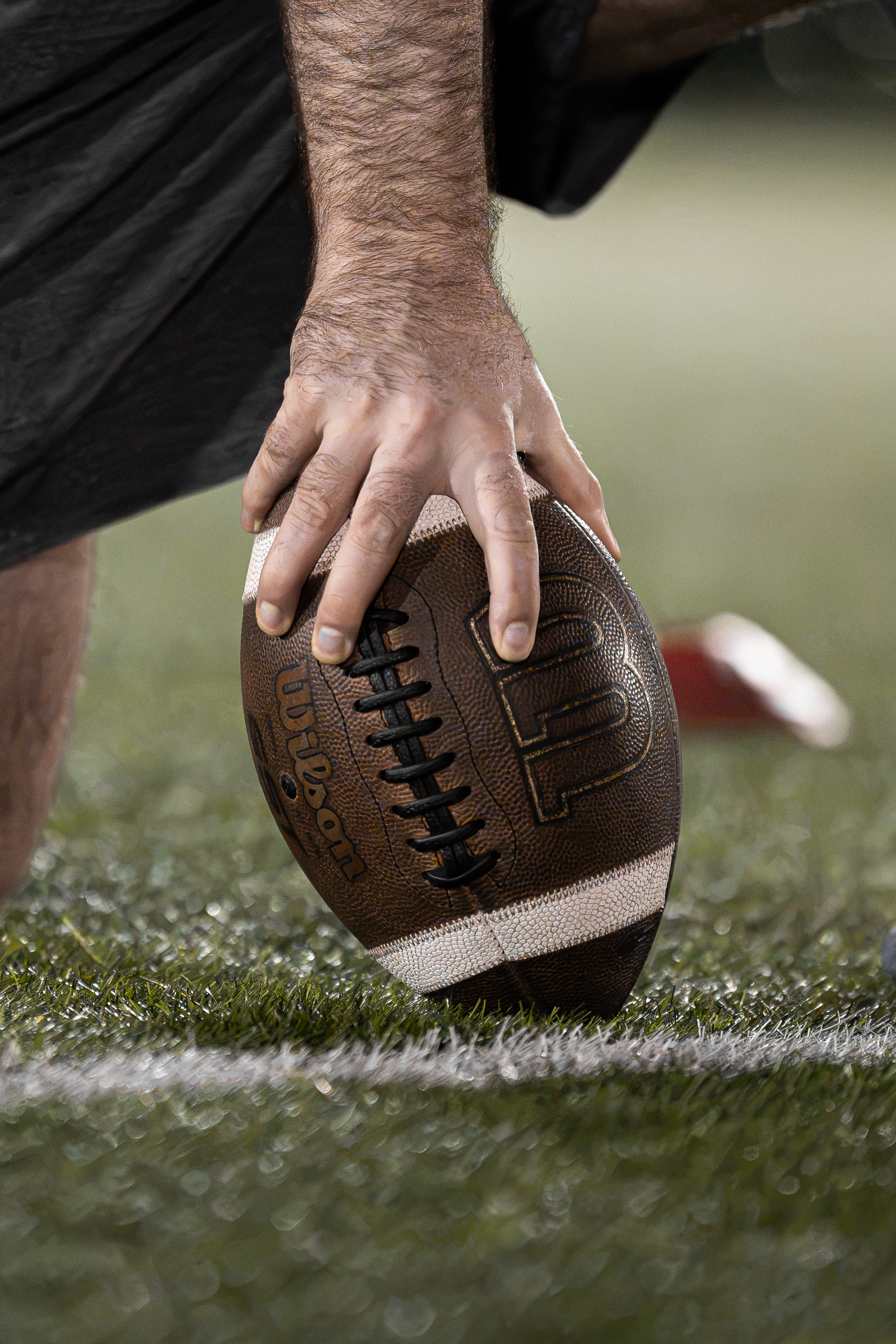 Close-up of a person's hand holding an American football on a grassy field, with part of their black shirt visible.