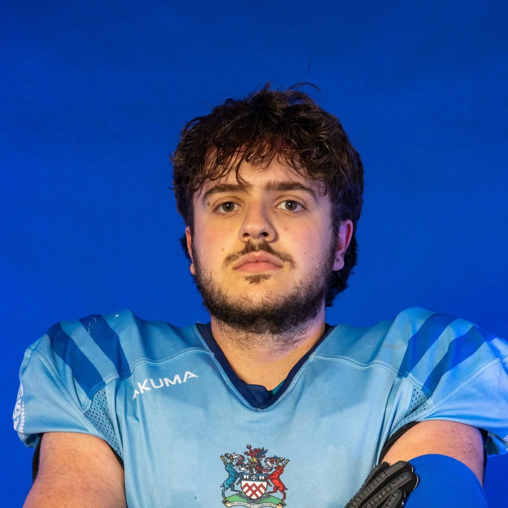 A football player in a blue team uniform holding a football, wearing a blue helmet with the Gloas logo, standing in front of a blue background with a Gloas logo.