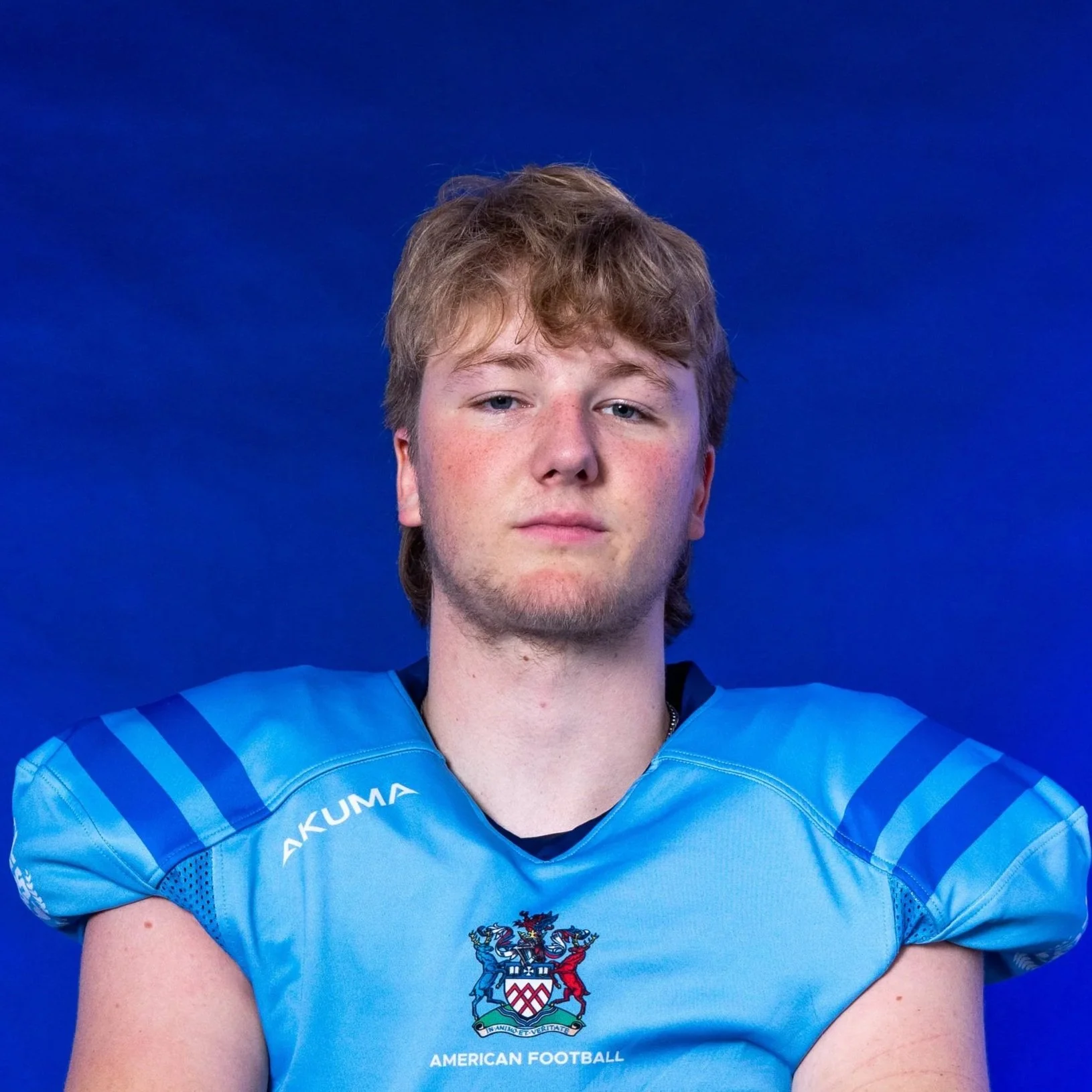 A football player in a light blue and black uniform holding a football, wearing a blue helmet with the Glaad logo, in front of a navy blue background with the Glaad logo and a small Michigan State Spartan logo.