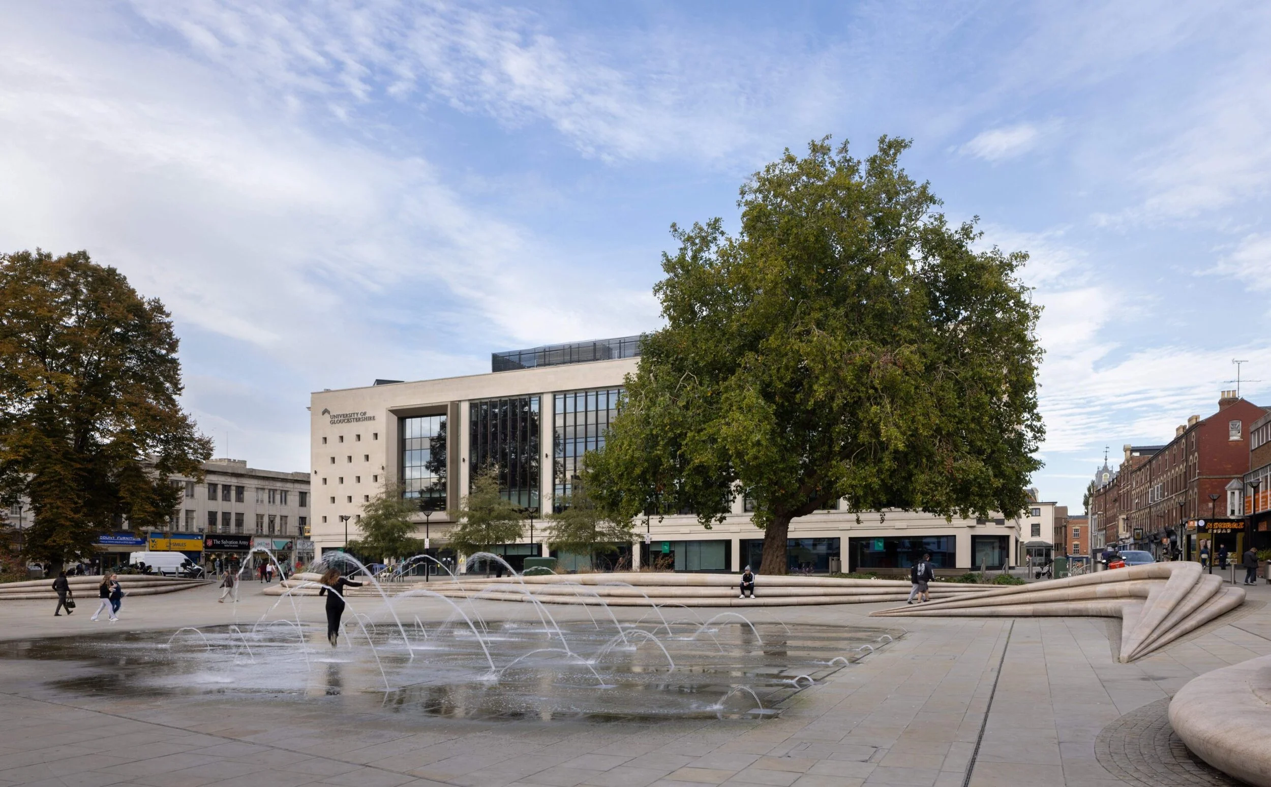 Public square with a modern white building labeled 'University of Gloucestershire,' large trees, a fountain with multiple streams of water, and people walking and sitting around.