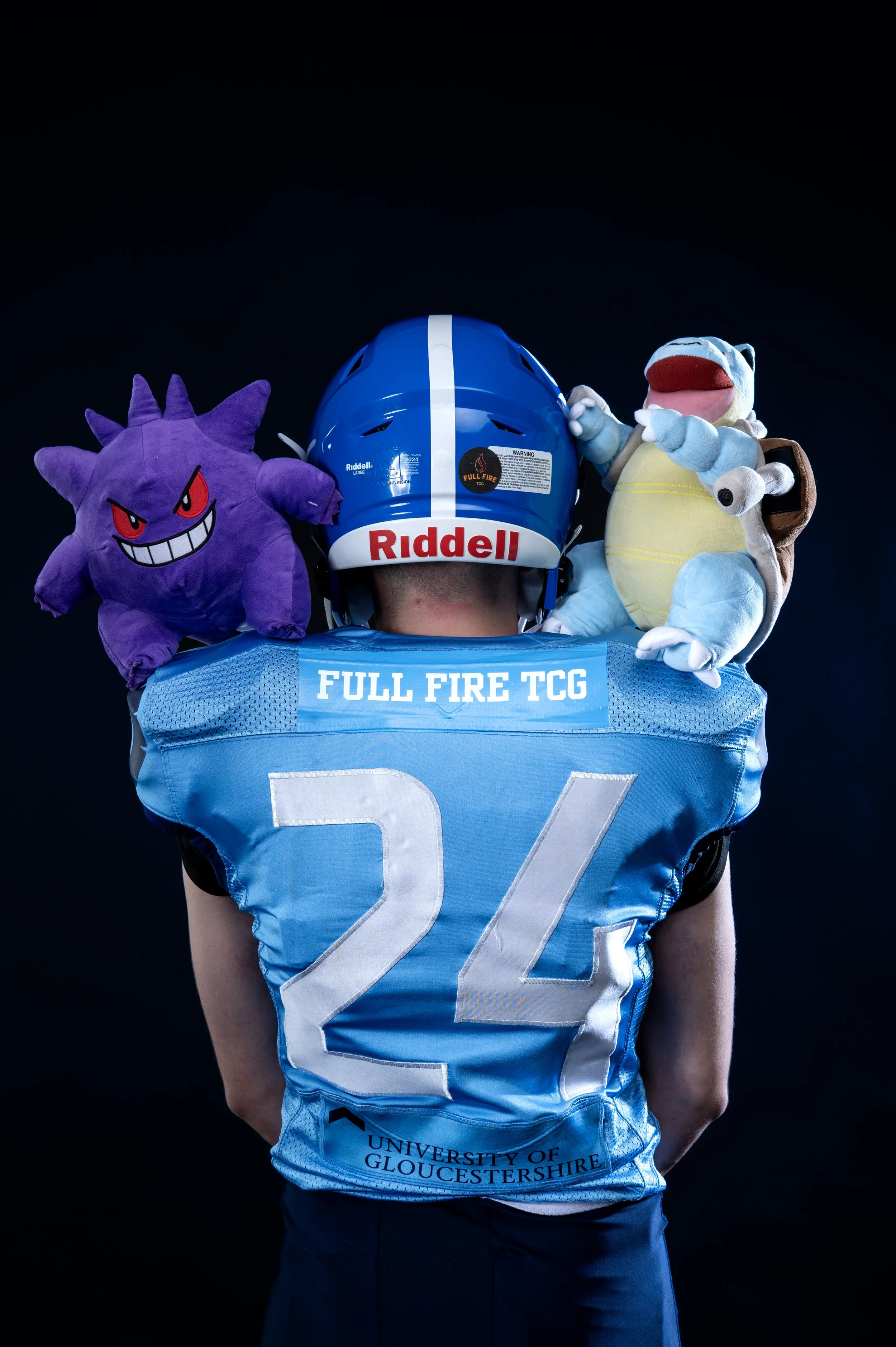 Back of a person wearing a blue football jersey with the number 22, a blue helmet with a white stripe and the word 'Riddell,' and a blue shoulder pad with 'Full Fire TCG' printed on it. They have plush toys of a purple Spiky Gengar and a blue and white Snorlax on their shoulders.