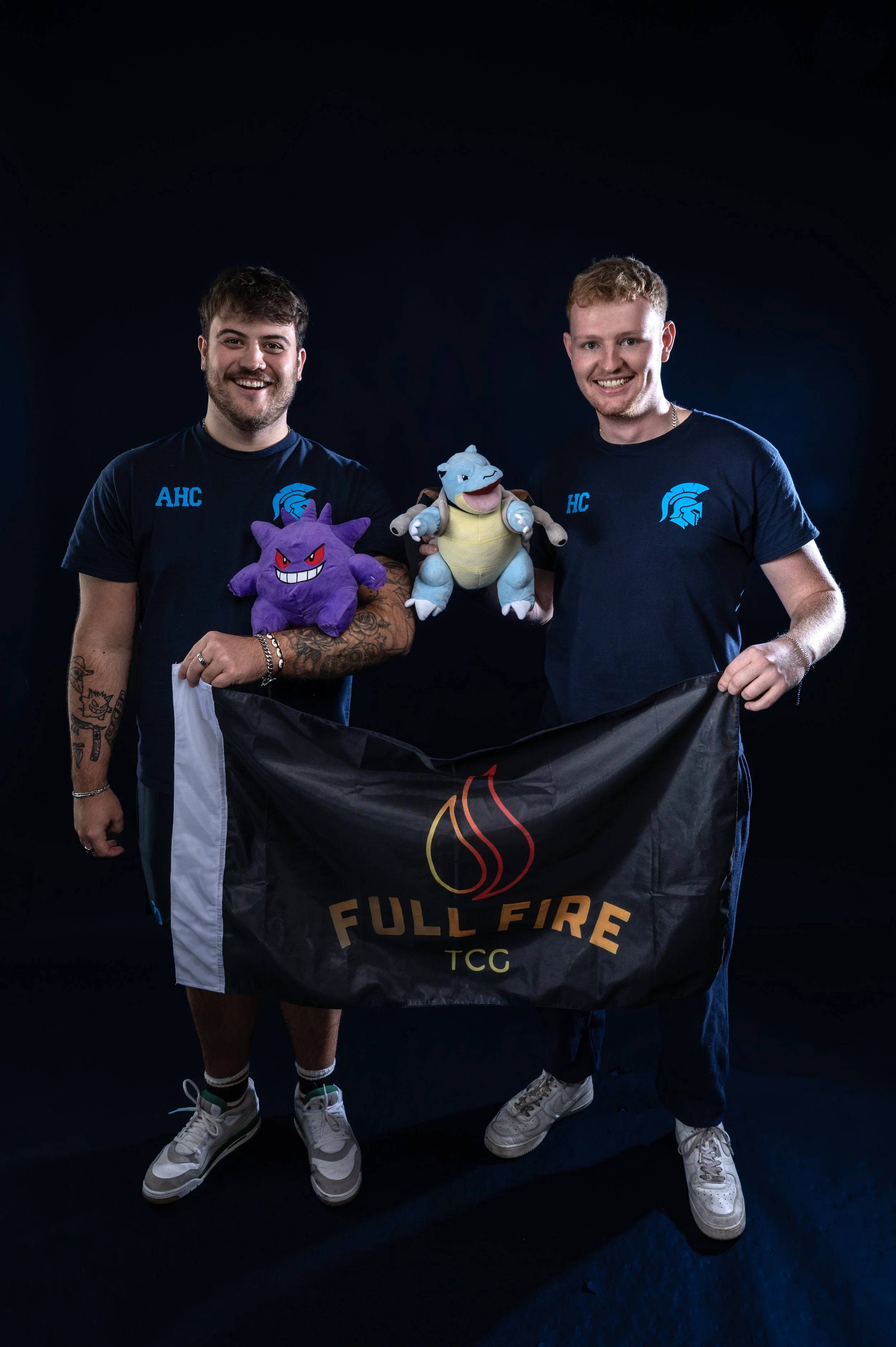Two young men standing side by side, smiling, wearing matching black t-shirts with blue logos, holding Pokémon plush toys of Gengar and Blastoise, and a black flag with the words "Full Fire" and a flame logo in front of them against a dark background.