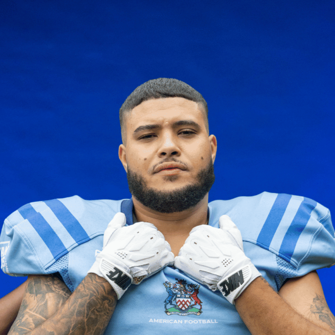 A football player in a blue and black uniform holding a football, wearing a blue helmet with the word "Glads" on it, standing in front of a dark blue background with the word "Glads" and a logo.