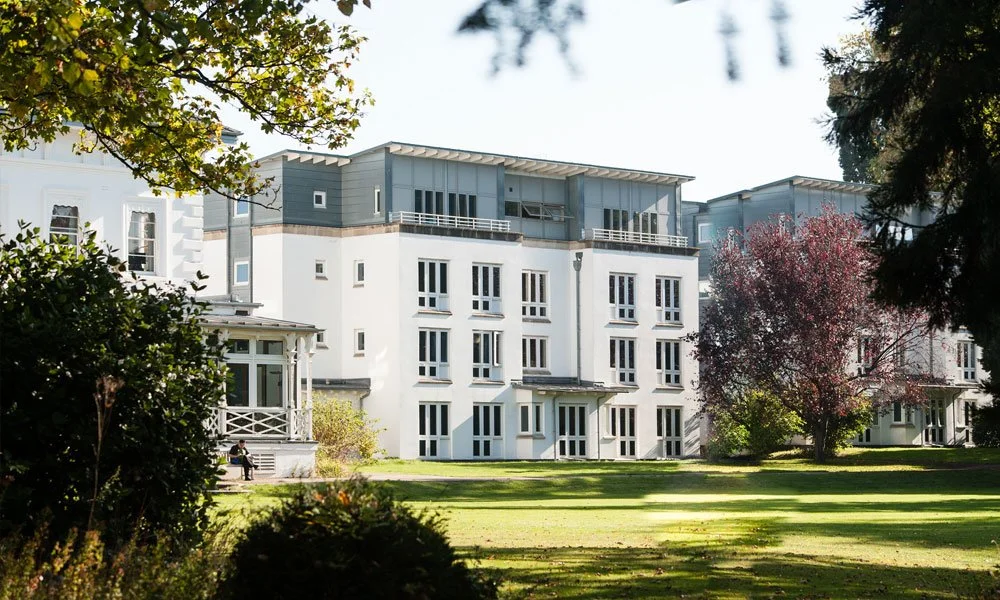 A modern multi-story white apartment building with large windows, situated in a lush green park with trees and well-maintained grass.