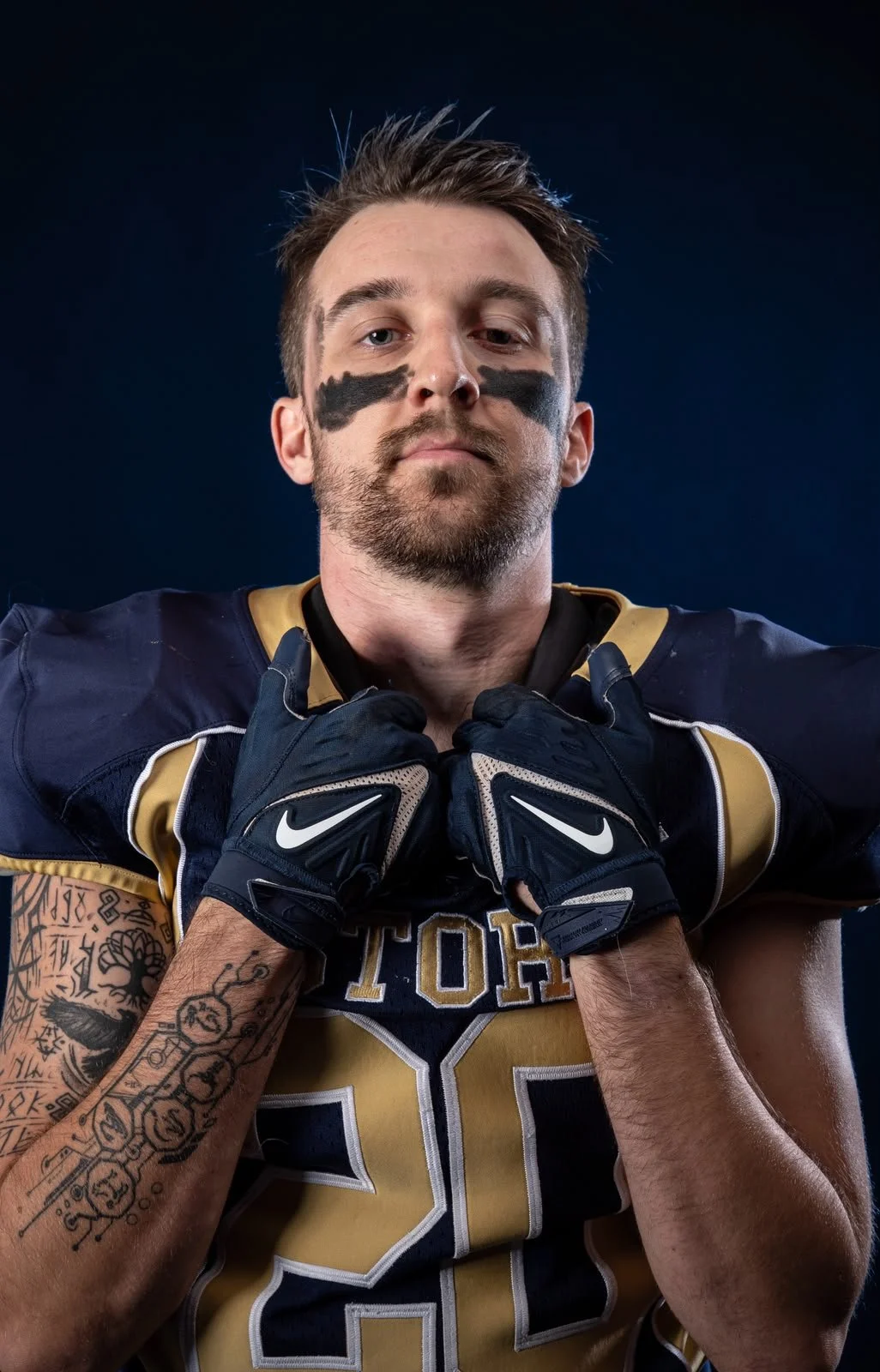 A young male football player in a navy and gold uniform, wearing black gloves, with painted black stripes under his eyes, holding his jersey collar with both hands, and displaying tattooed arms, against a dark background.