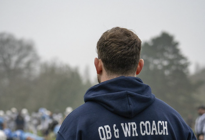 Back of a man in a navy hoodie with 'QB & WR COACH' printed on it, overlooking a football practice or game field with players and coaches in the background.