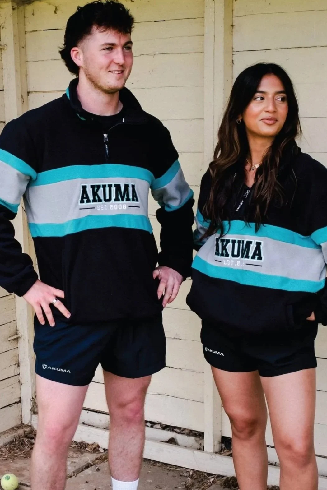 Two people wearing black and blue sports jackets with the word "AKUMA" printed on them, standing outdoors near a wooden fence. The man has short dark hair and is wearing black shorts, while the woman has long dark hair and is also wearing black shorts. Both appear to be posing casually.