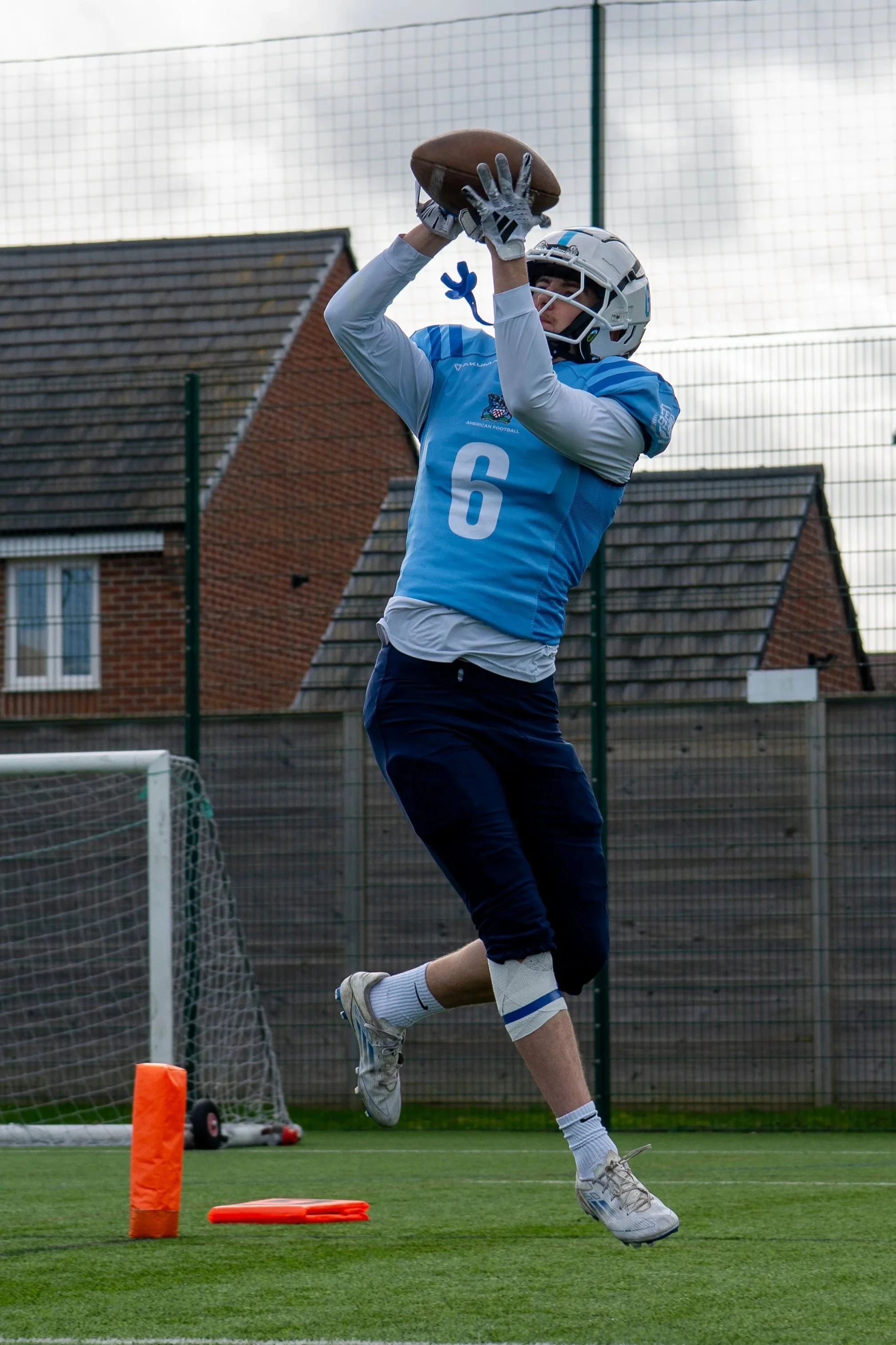 A young male football player wearing a white helmet, blue jersey with the number 6, and dark pants is jumping to catch a football during practice on a football field. There is a small orange cone and some equipment on the ground near him, with houses and a wire fence in the background.