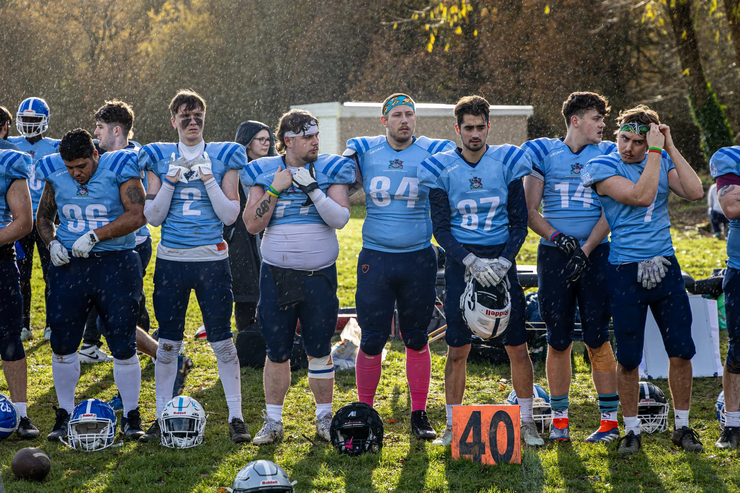 A group of young football players in blue uniforms standing on a grassy field, some with helmets on the ground in front of them. They are lined up, with some looking down or to the side, and appear to be in a moment of reflection or rest during a game or practice. There is an orange yard marker with the number 40 in front of them.