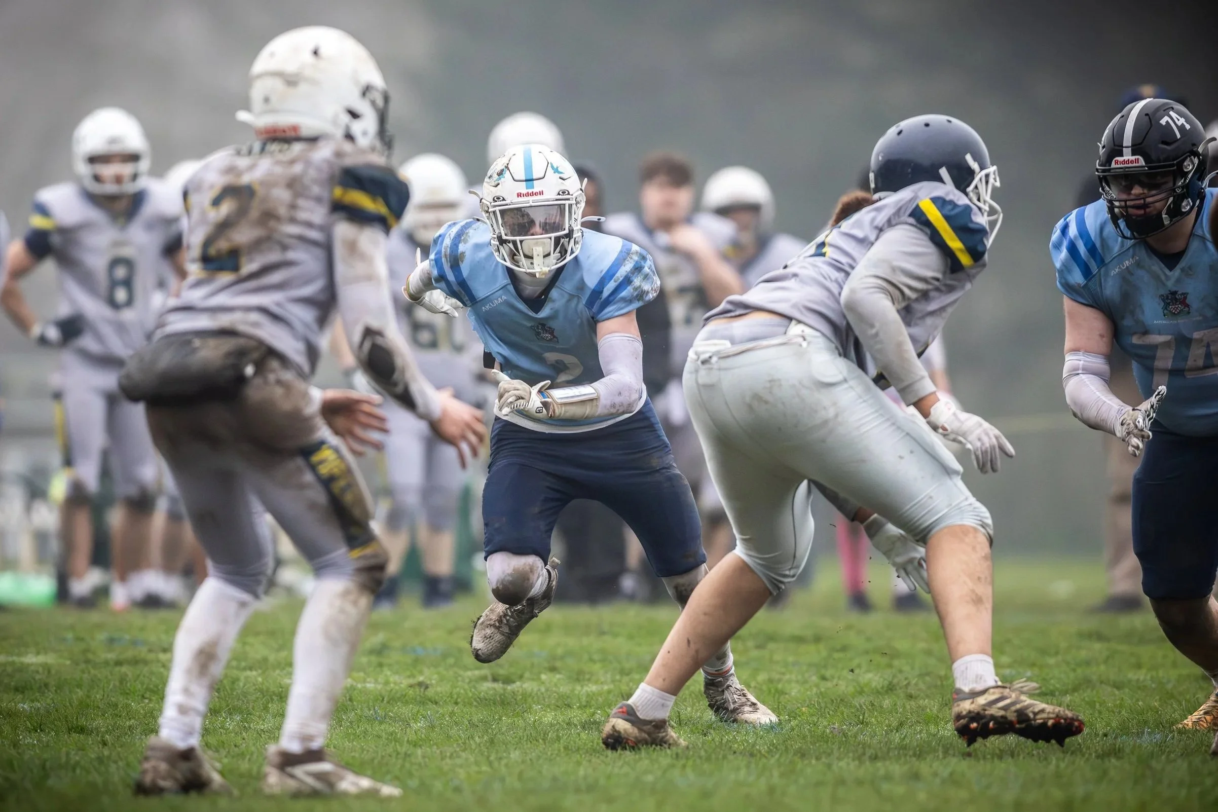 Youth football players in action during a game, wearing helmets and uniforms, on a grassy field with a foggy background.