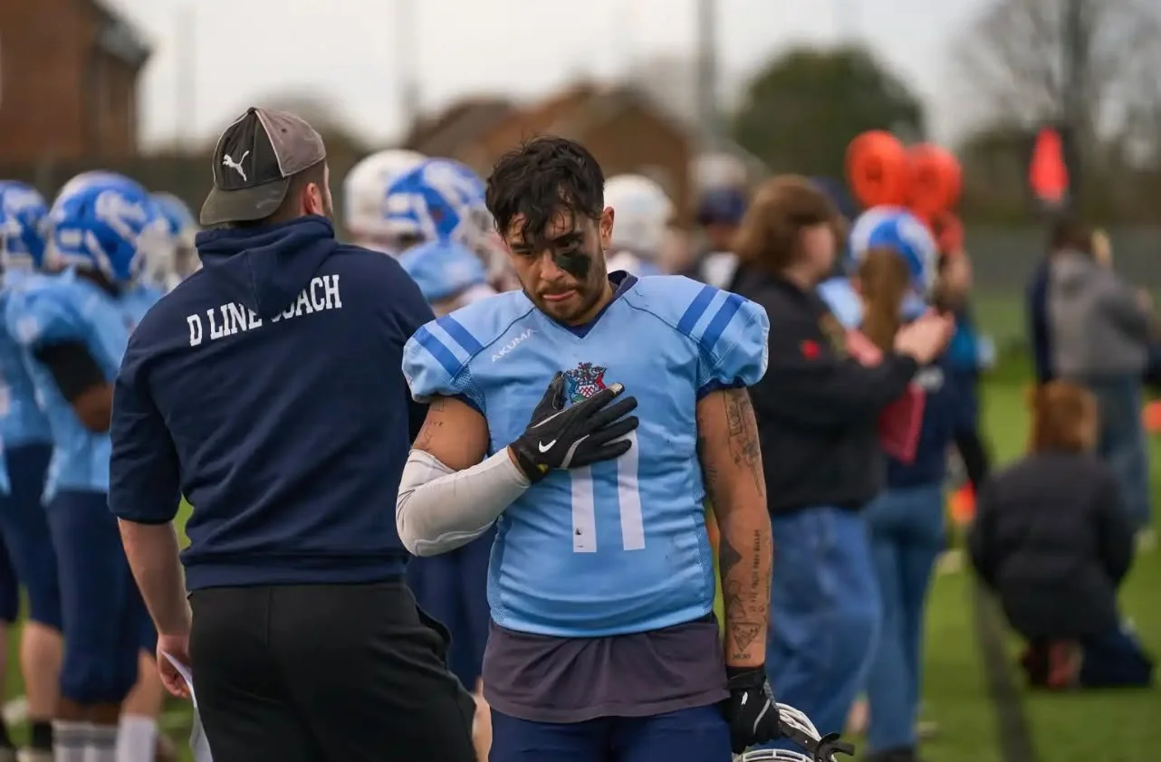 A football player in a blue jersey with face paint, standing on the field with his hand over his heart, with teammates and coaches in the background.