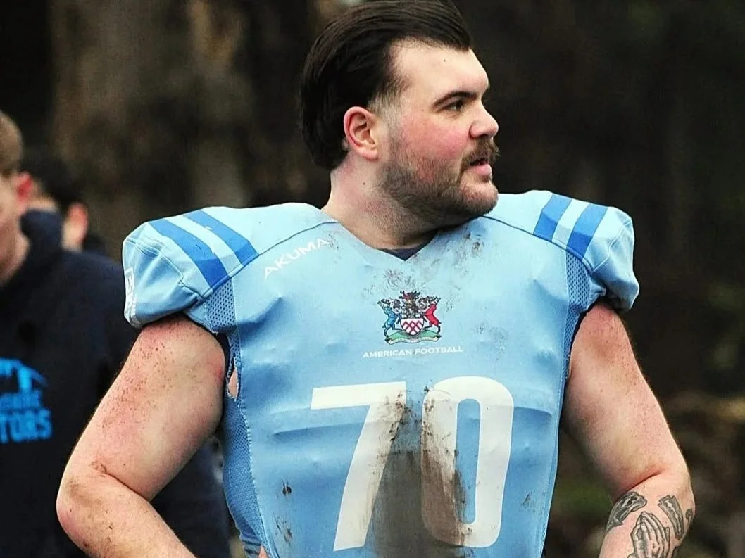 Man in a light blue American football uniform with the number 70, surrounded by mud, with dark hair and a beard, outdoors after a game.