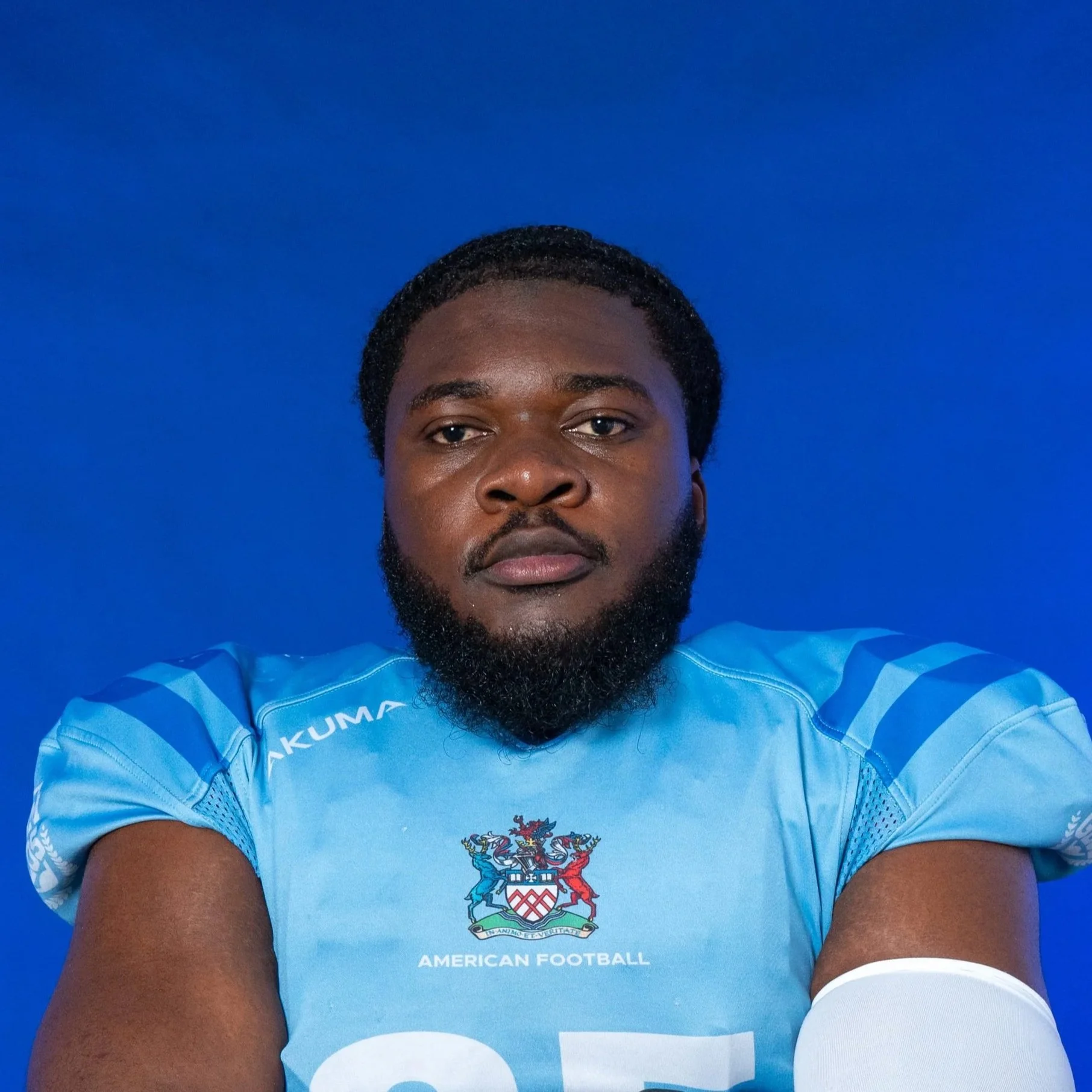 A football player in a blue GLOMS helmet and team jersey holding a football, standing against a GLOMS backdrop.